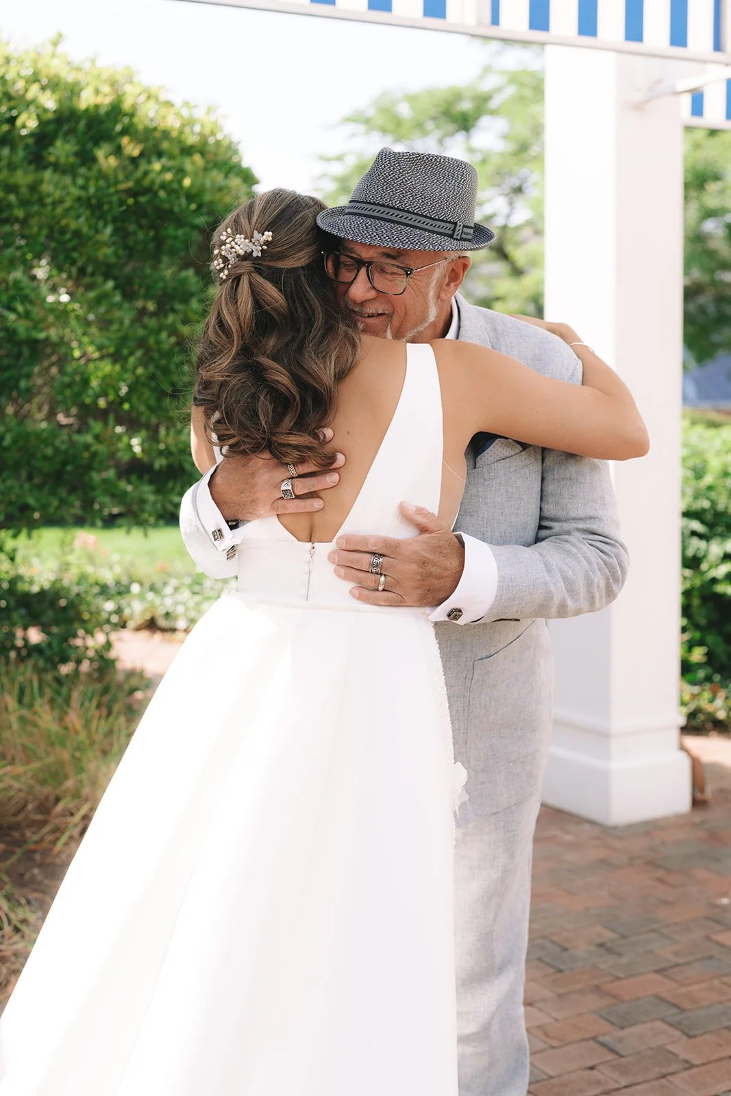A bride in a white wedding dress hugging an older man in a gray suit and hat outdoors.