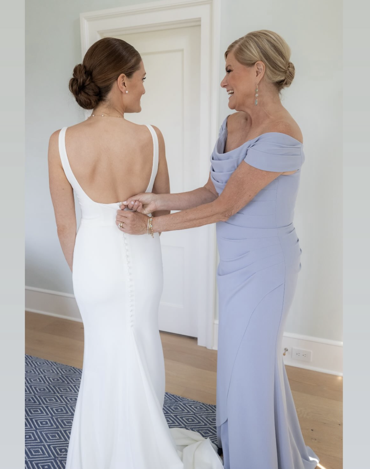 A bride in a white wedding dress standing with her back facing the camera, while an older woman in a light blue dress, likely her mother, adjusts the back of her dress and smiles.