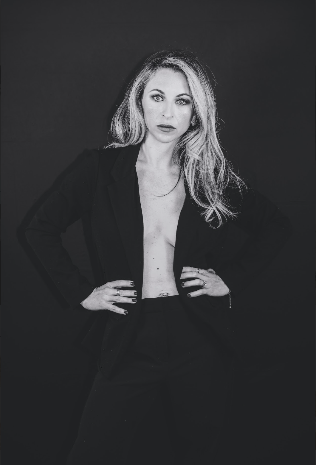 A woman with long, wavy blonde hair posing with her hands on her hips against a dark background. She is wearing a black blazer and pants, with a no shirt or top underneath, exposing her chest and abdomen. The photo is in black and white.