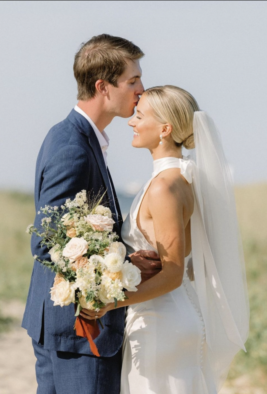 A wedding couple is on a beach, with the groom kissing the bride on her forehead. The bride is holding a large bouquet of flowers and is wearing a white dress and veil.