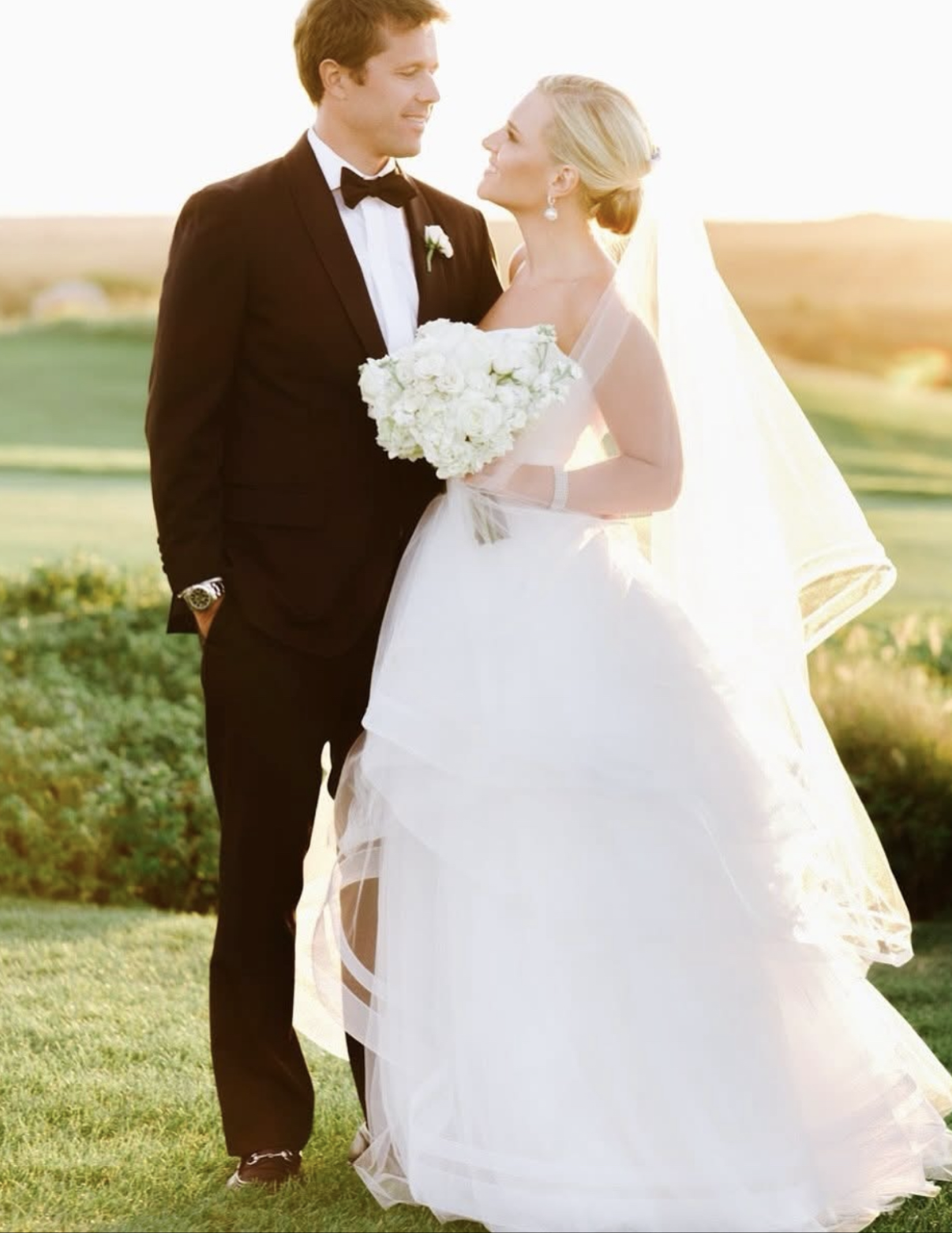 Bride and groom standing outdoors during sunset, wearing wedding attire, with the bride holding a bouquet of white flowers.