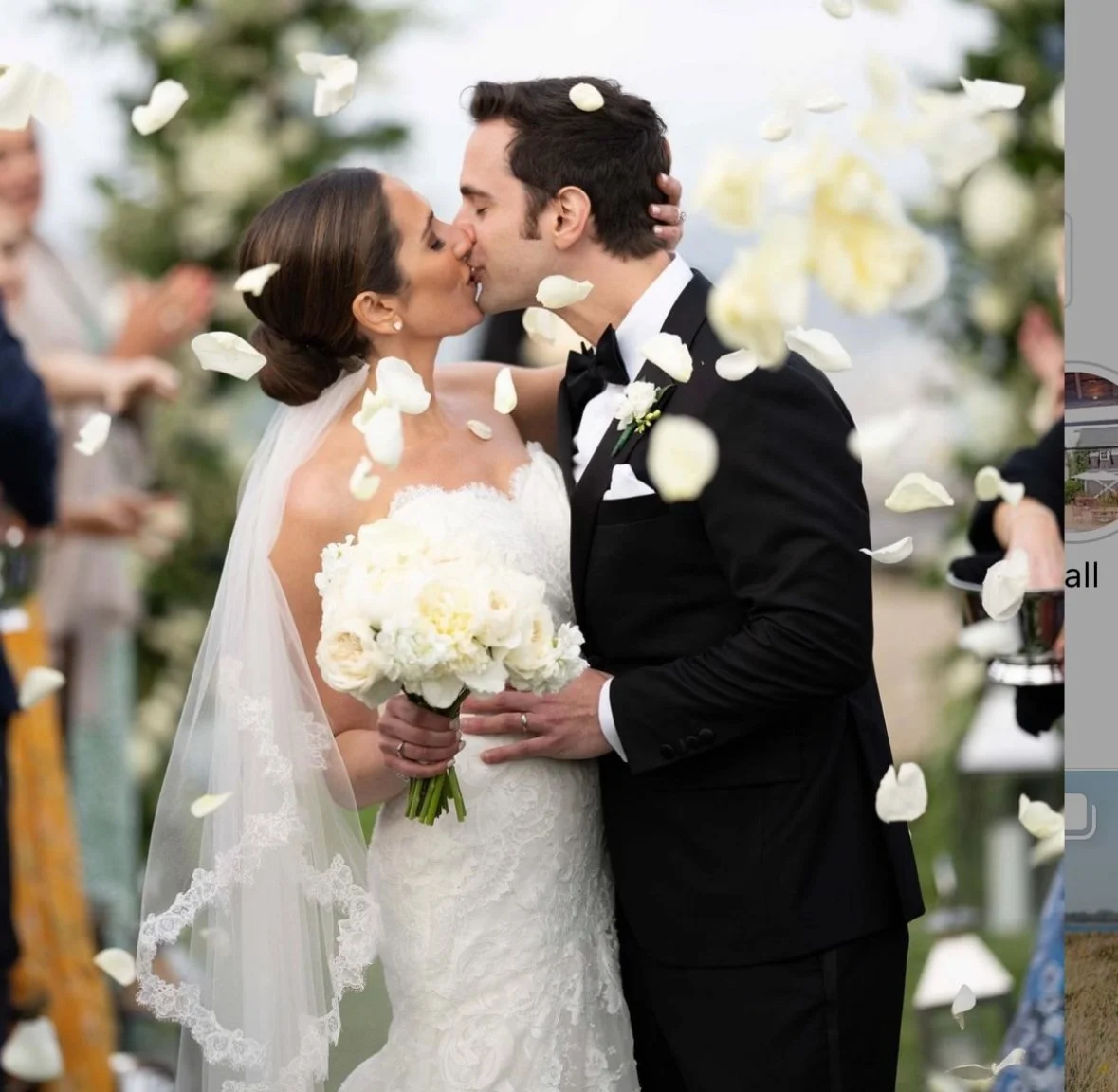 A bride and groom kiss during their wedding ceremony, surrounded by falling flower petals, with guests in the background.
