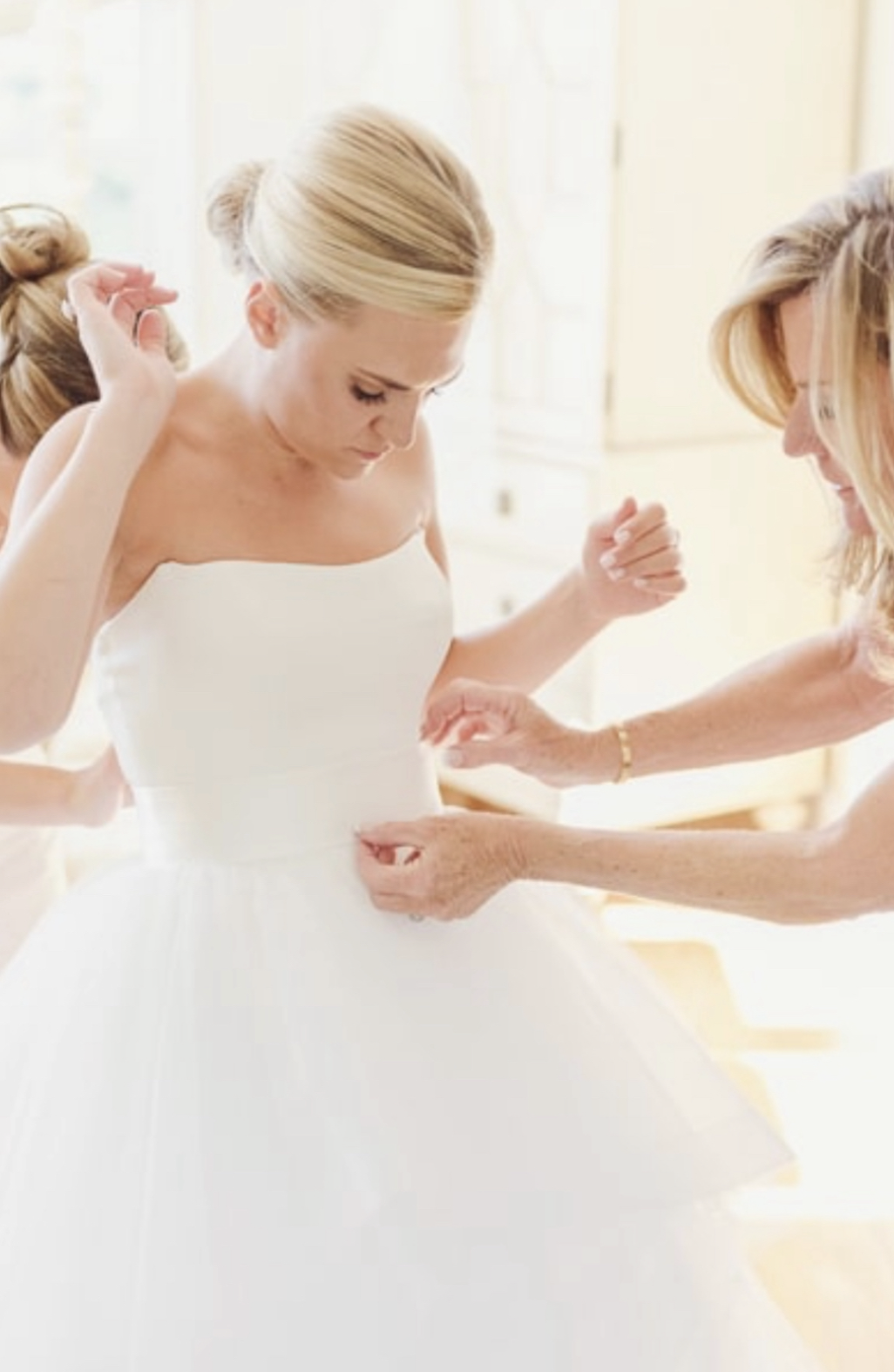 A bride in a white wedding dress is being assisted by older women, likely her mother or close family, with adjusting her dress before the wedding ceremony.