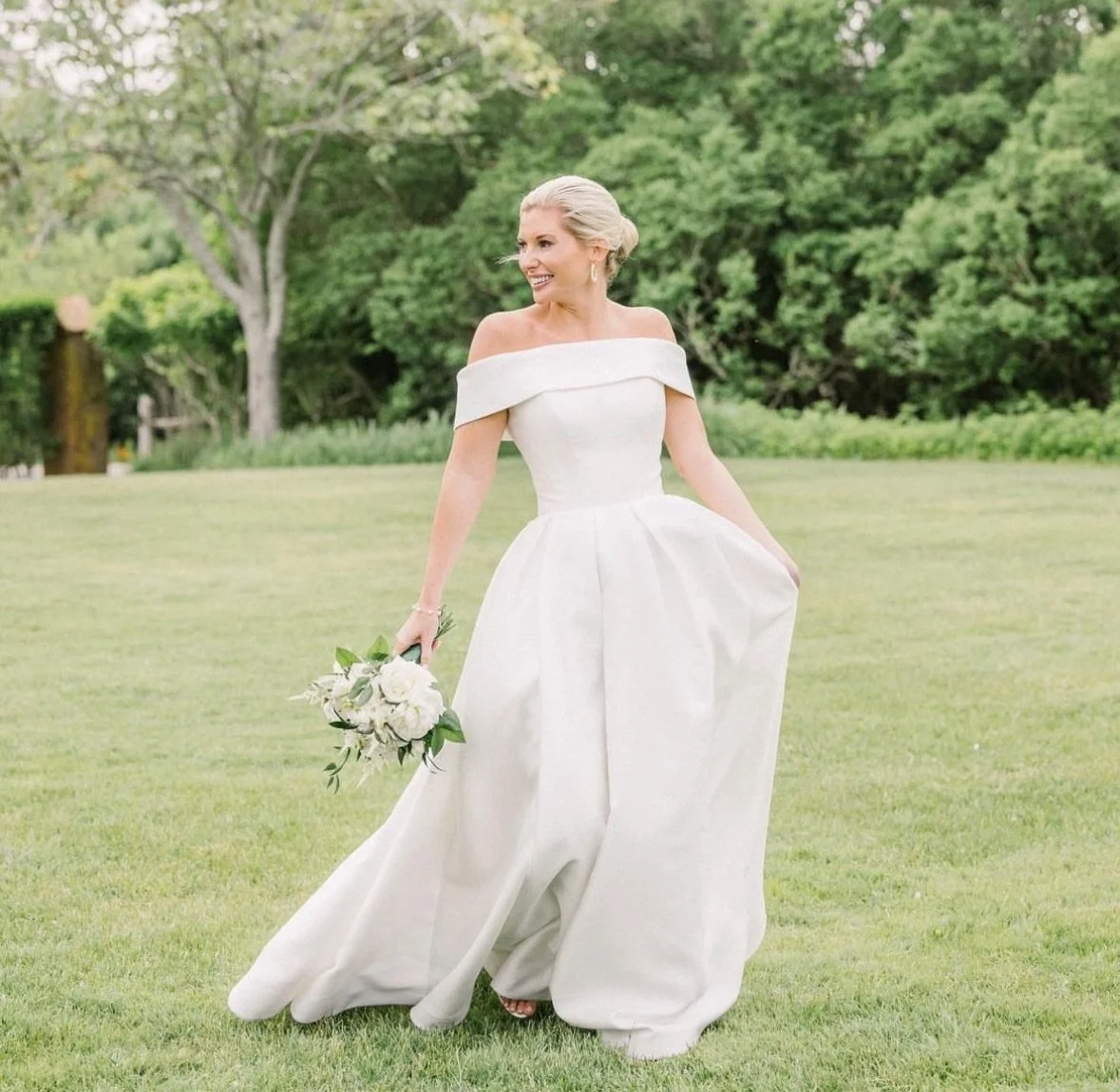 A woman in a white off-the-shoulder wedding dress holding a bouquet of white flowers, outdoors on a grassy field with trees in the background.