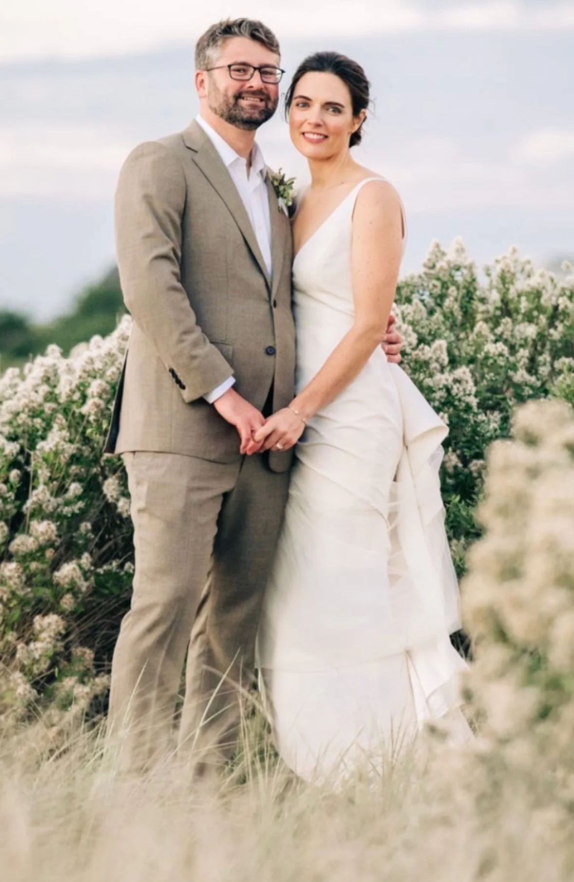 A newlywed couple stands outdoors on their wedding day, holding hands and smiling, with white flowers and greenery in the background.