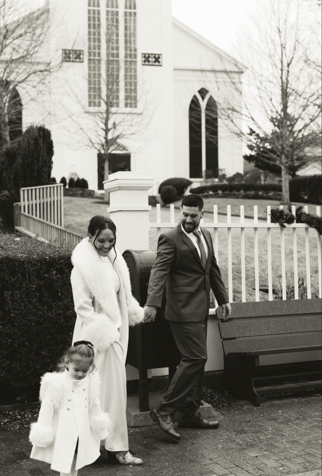 A black-and-white photo of a happy couple holding hands, with a young girl walking in front of them, near a white church with tall windows and autumn trees in the background.