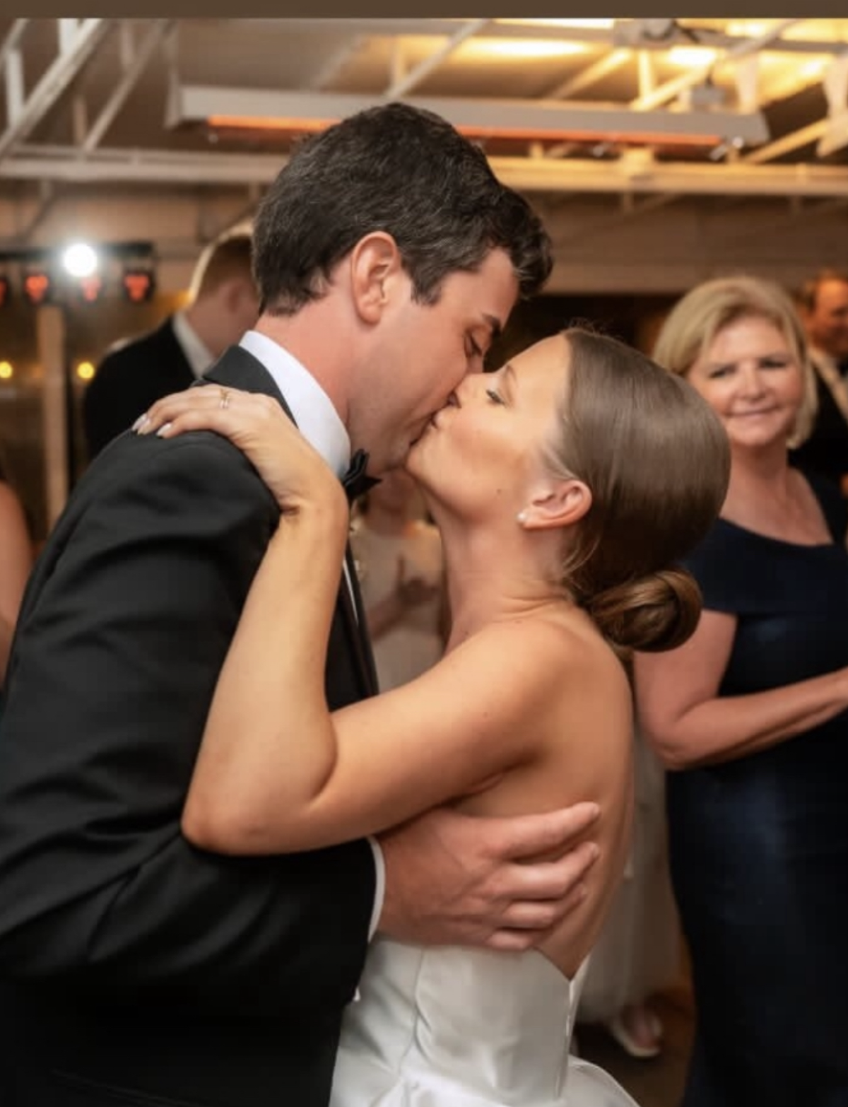 A newlywed couple sharing a passionate kiss at their wedding reception, with guests in the background.