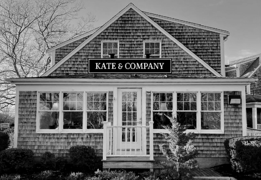 A two-story house with shingle siding, large front windows, a small front porch with a white railing, and a sign reading 'KATE & COMPANY' on the upper part of the house.