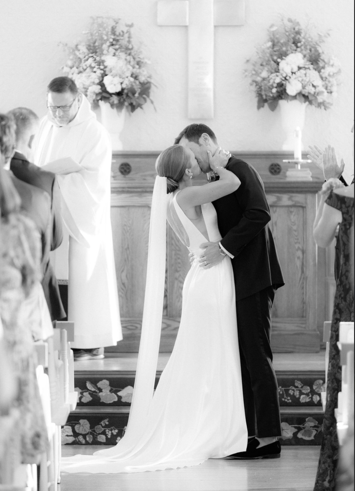 A black-and-white photo of a wedding ceremony showing a bride and groom kissing at the altar. The bride wears a white dress with a long train and veil, while the groom wears a black tuxedo. An officiant dressed in white stands nearby, and guests are 