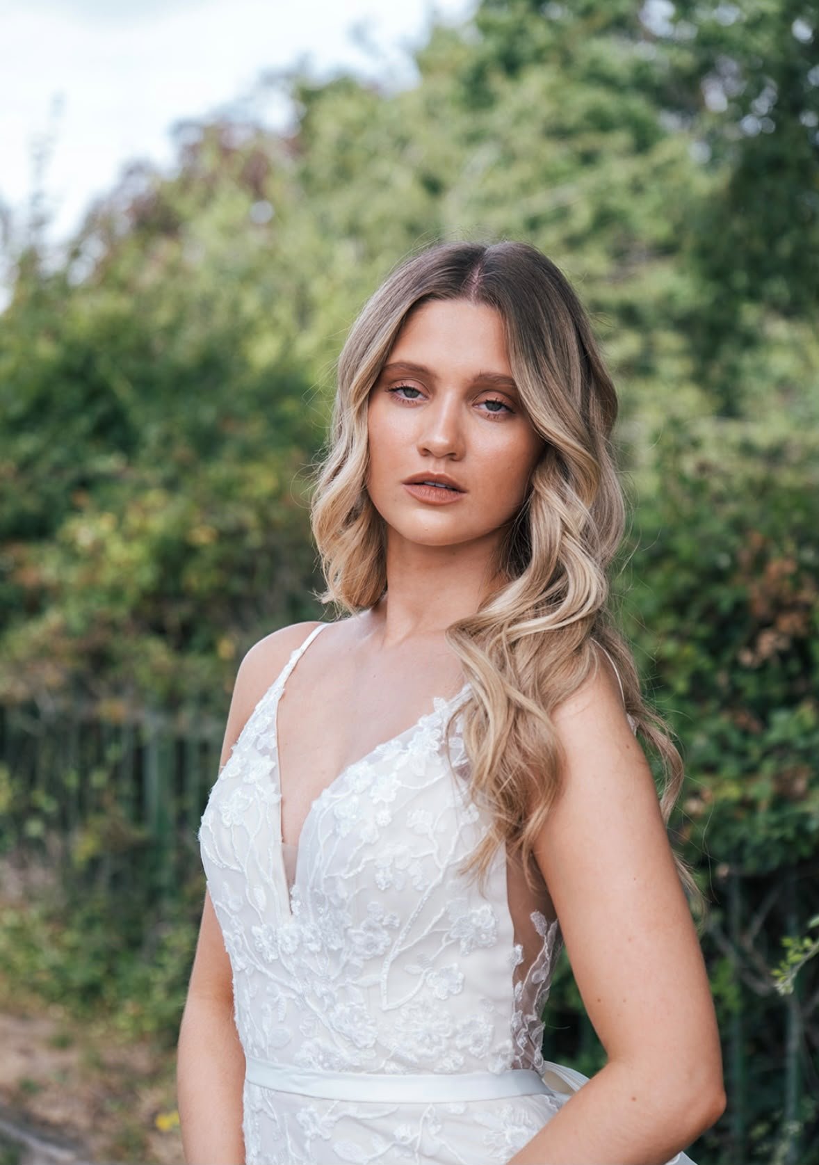 A young woman with wavy blonde hair in a white lace dress standing outdoors with green trees in the background.