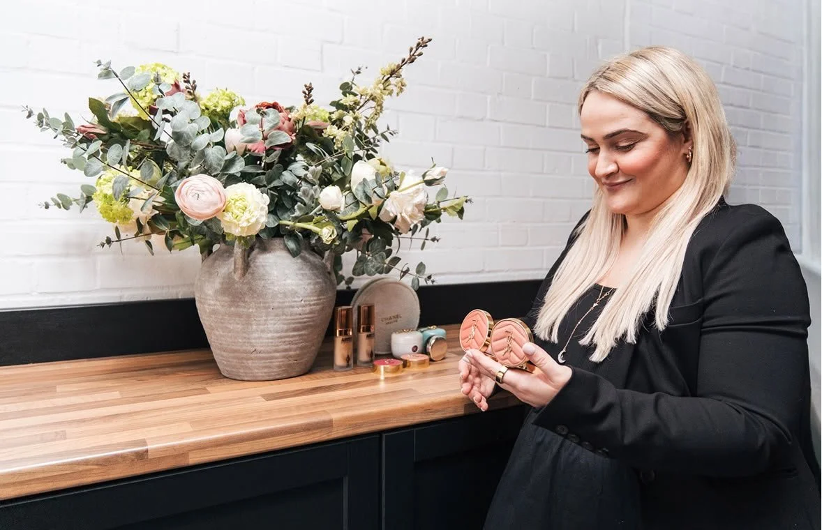 A woman with long blonde hair wearing a black blazer holding makeup compacts in a room with a bouquet of flowers on a wooden counter.