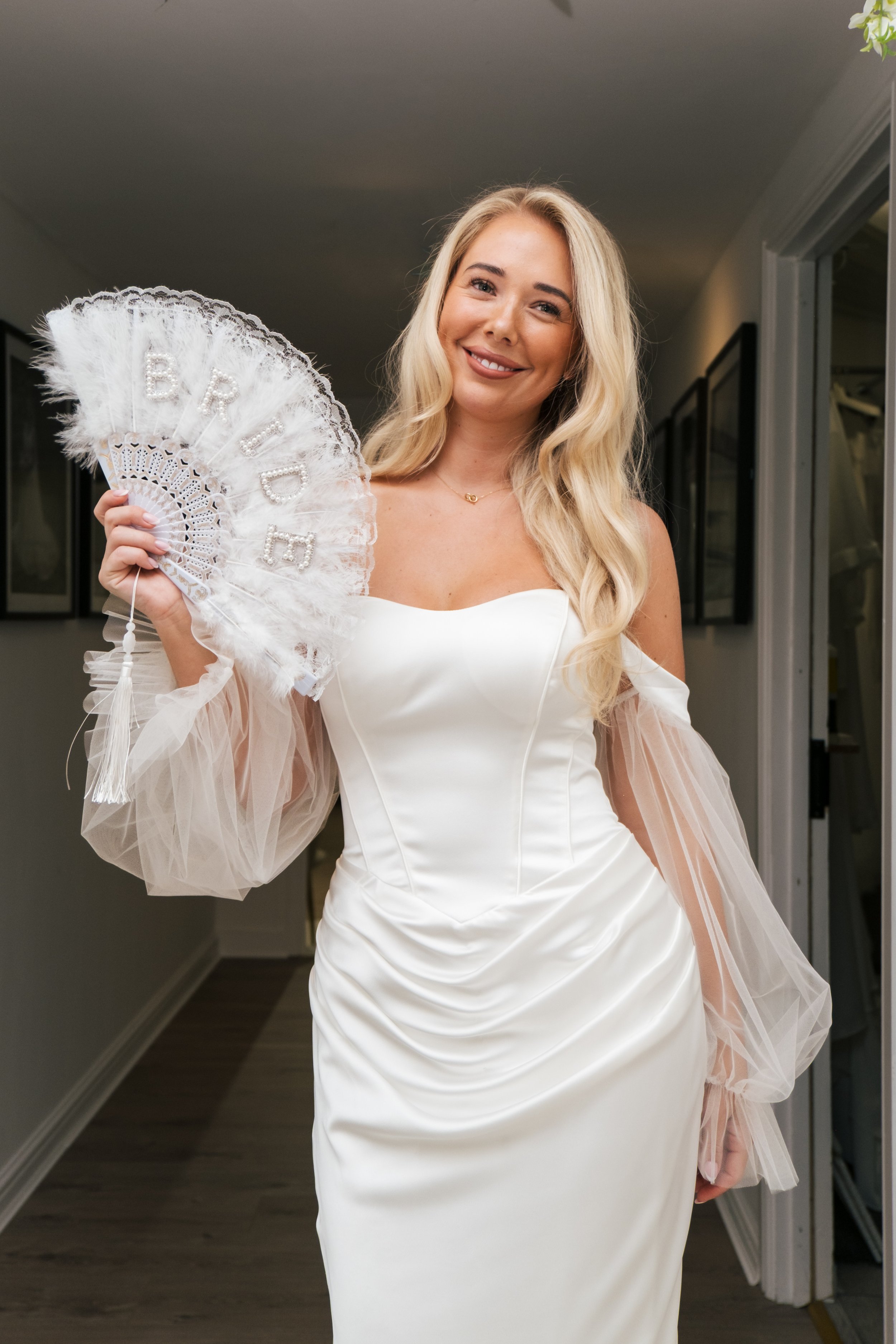 A woman with long blonde hair smiling and holding a white lace fan in a white off-shoulder dress with sheer long sleeves, standing in a hallway with framed pictures on the wall.