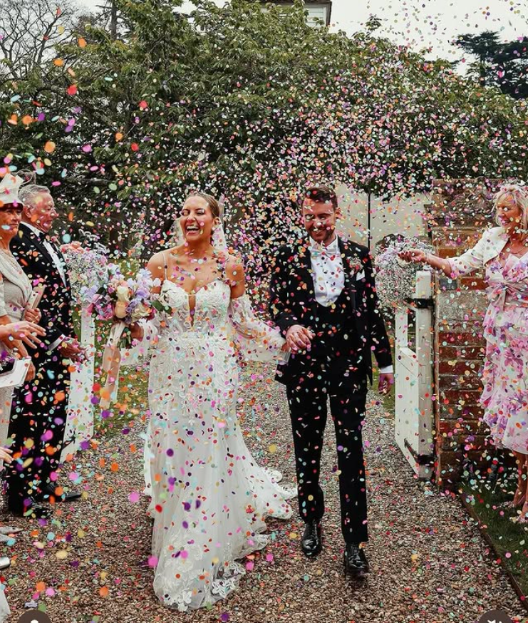 Couple in wedding attire walking hand-in-hand under shower of colorful confetti, with smiling guests on either side in a garden setting.