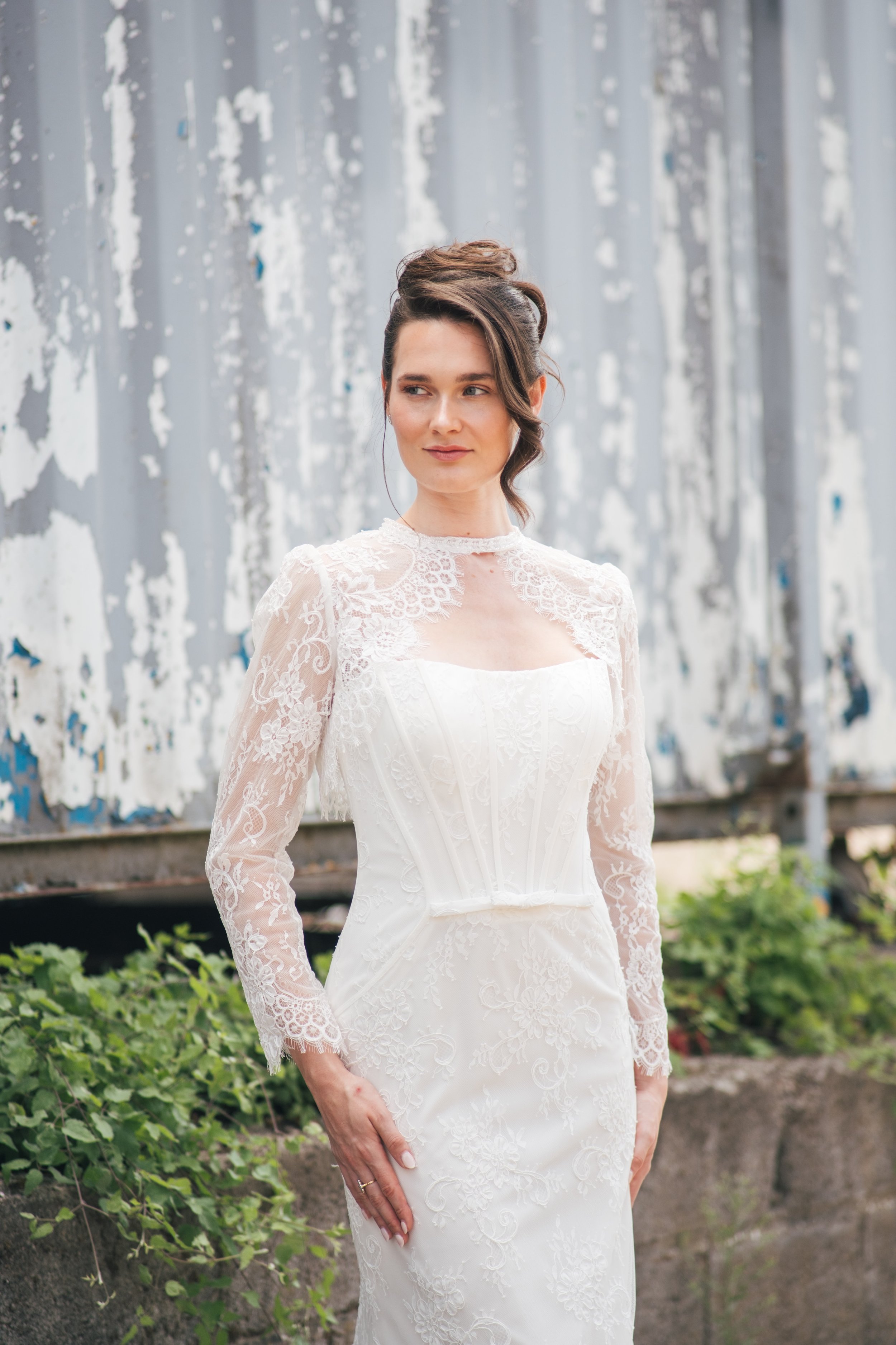 A woman wearing a white lace wedding dress standing outdoors near green plants and a weathered metal wall.