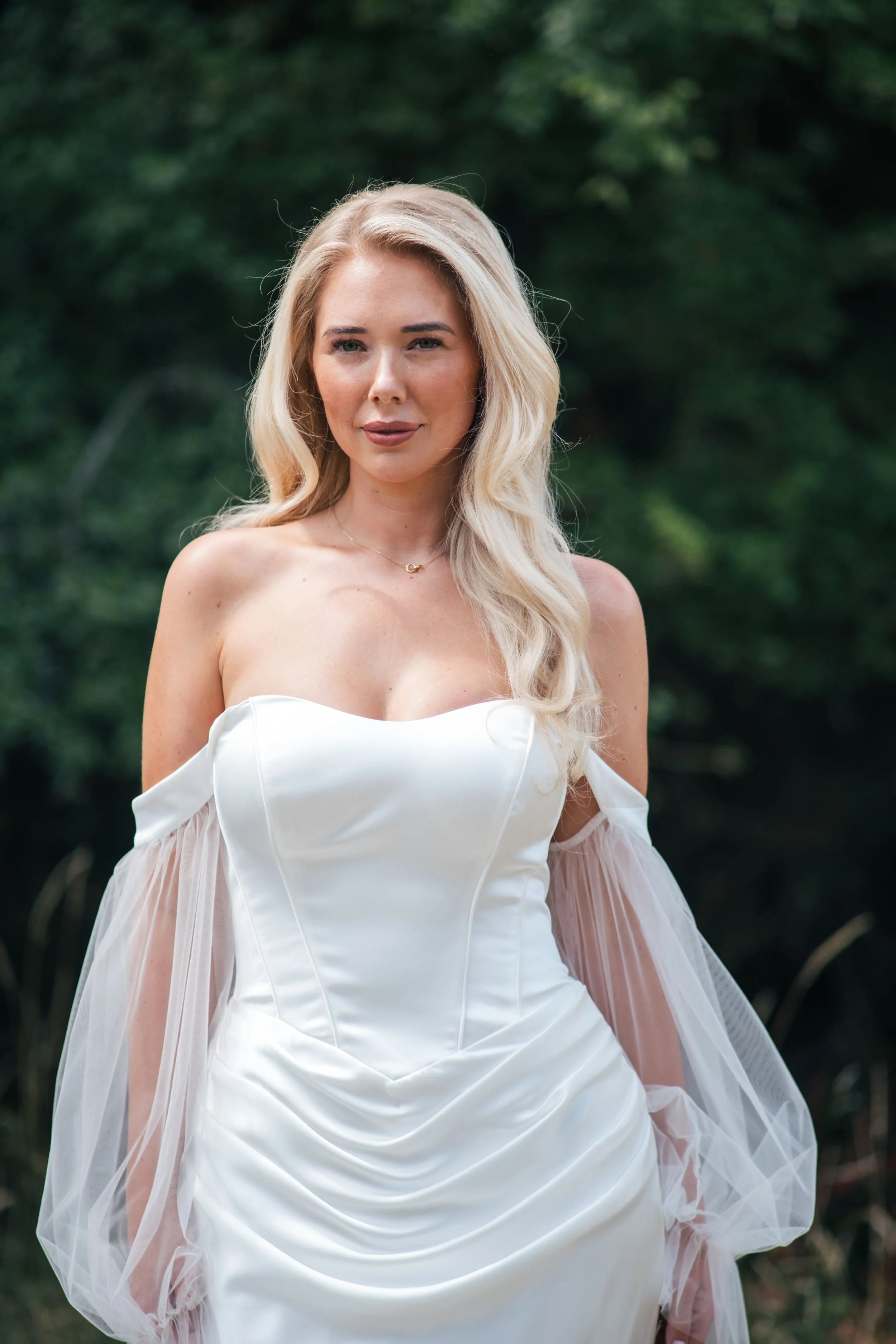 A woman in a strapless white dress standing outdoors with a blurred green background.
