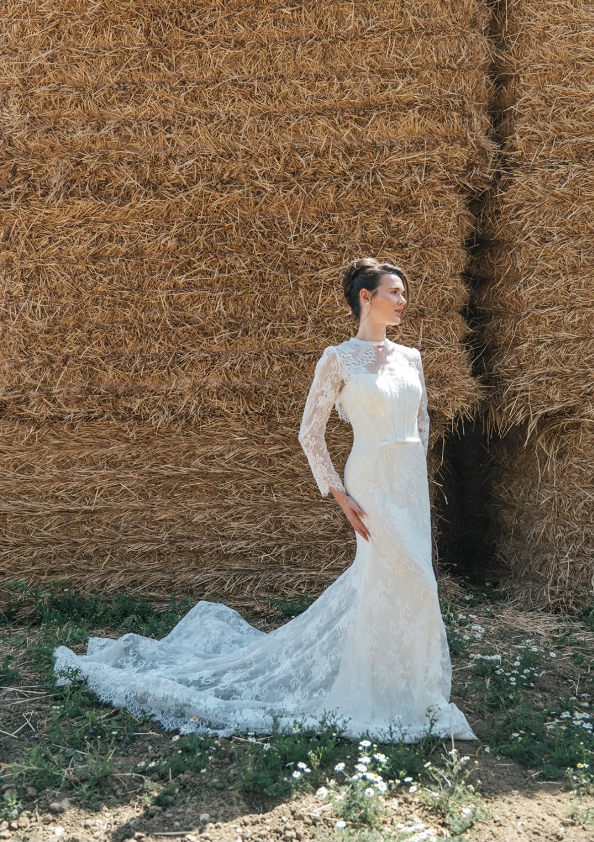 A woman in a white lace wedding dress stands outdoors in front of large hay bales, with small white flowers at her feet and greenery nearby.