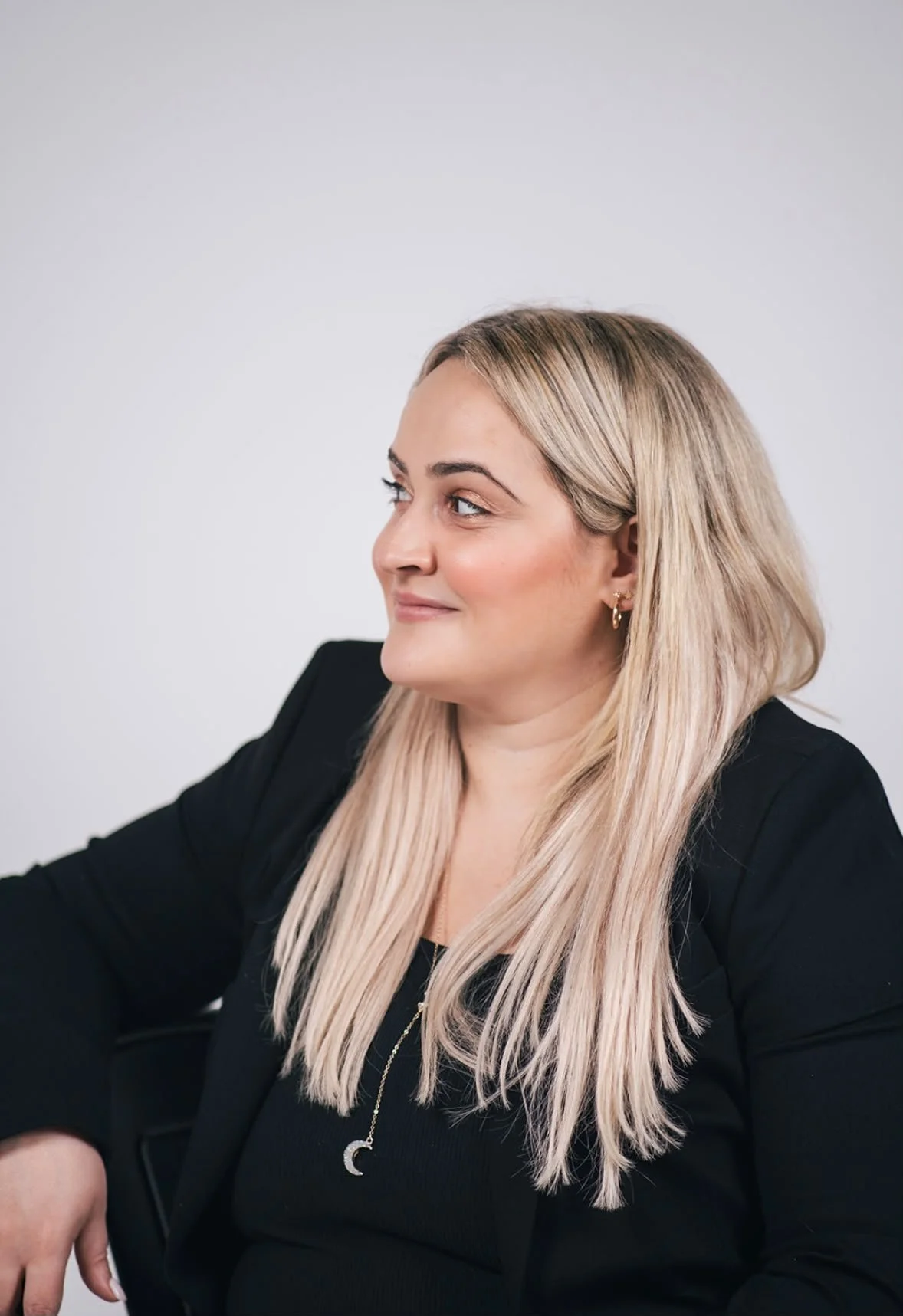 A woman with long blonde hair, wearing a black blazer, and gold jewelry, sitting against a plain gray background.