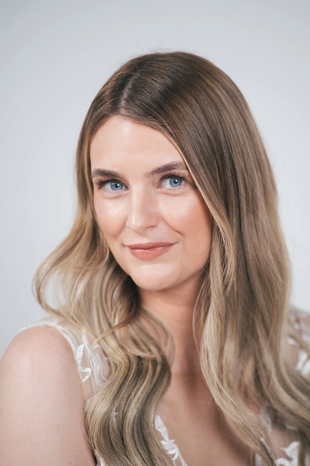 Close-up portrait of a woman with long wavy blonde hair, blue eyes, and a slight smile, wearing a white lace top, against a plain light background.