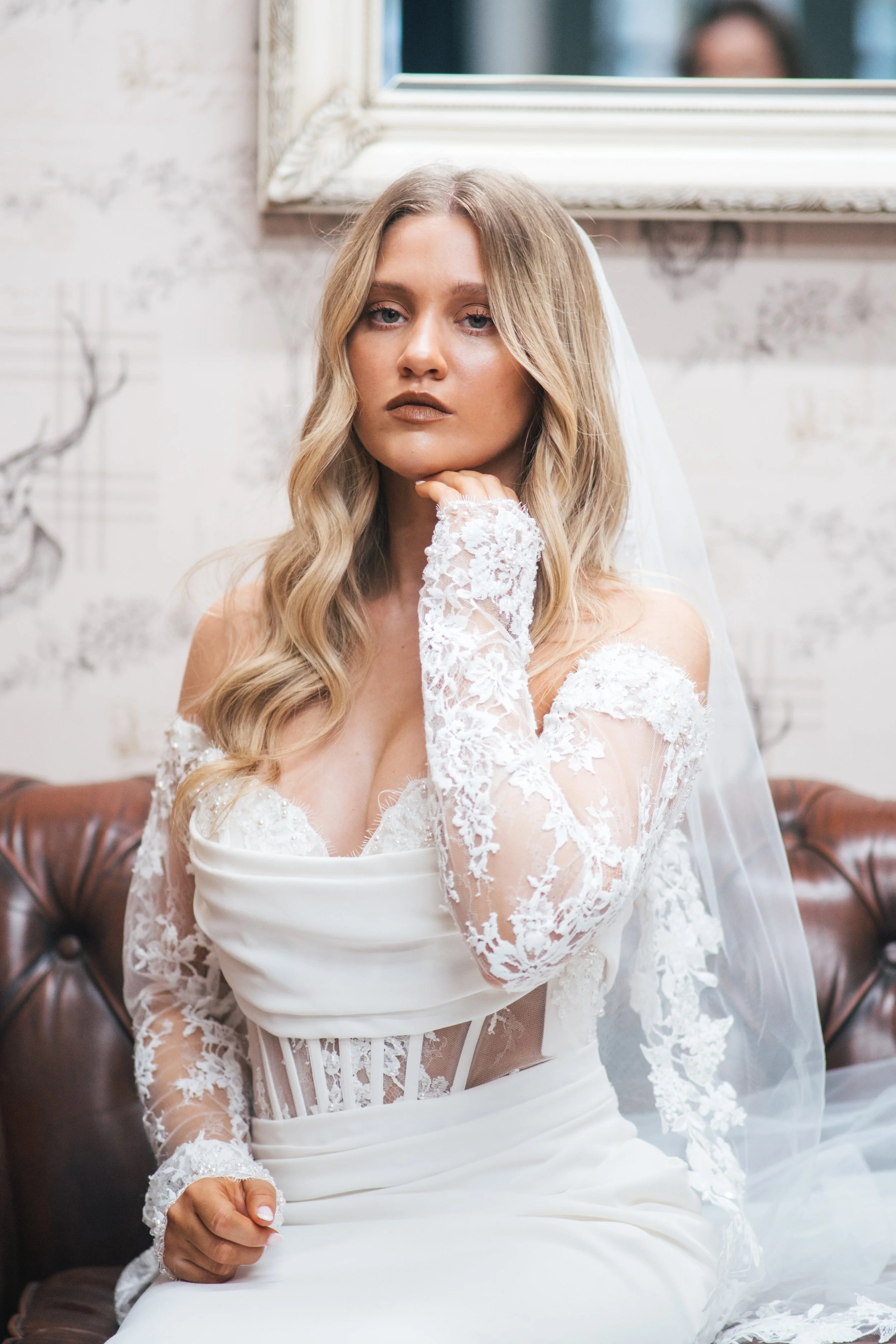 A bride sitting on a leather sofa, wearing a white lace wedding dress with sheer lace long sleeves and a veil, with her hair styled in loose waves. She rests her chin on her hand, looking directly at the camera, with a mirror reflecting a blurred per