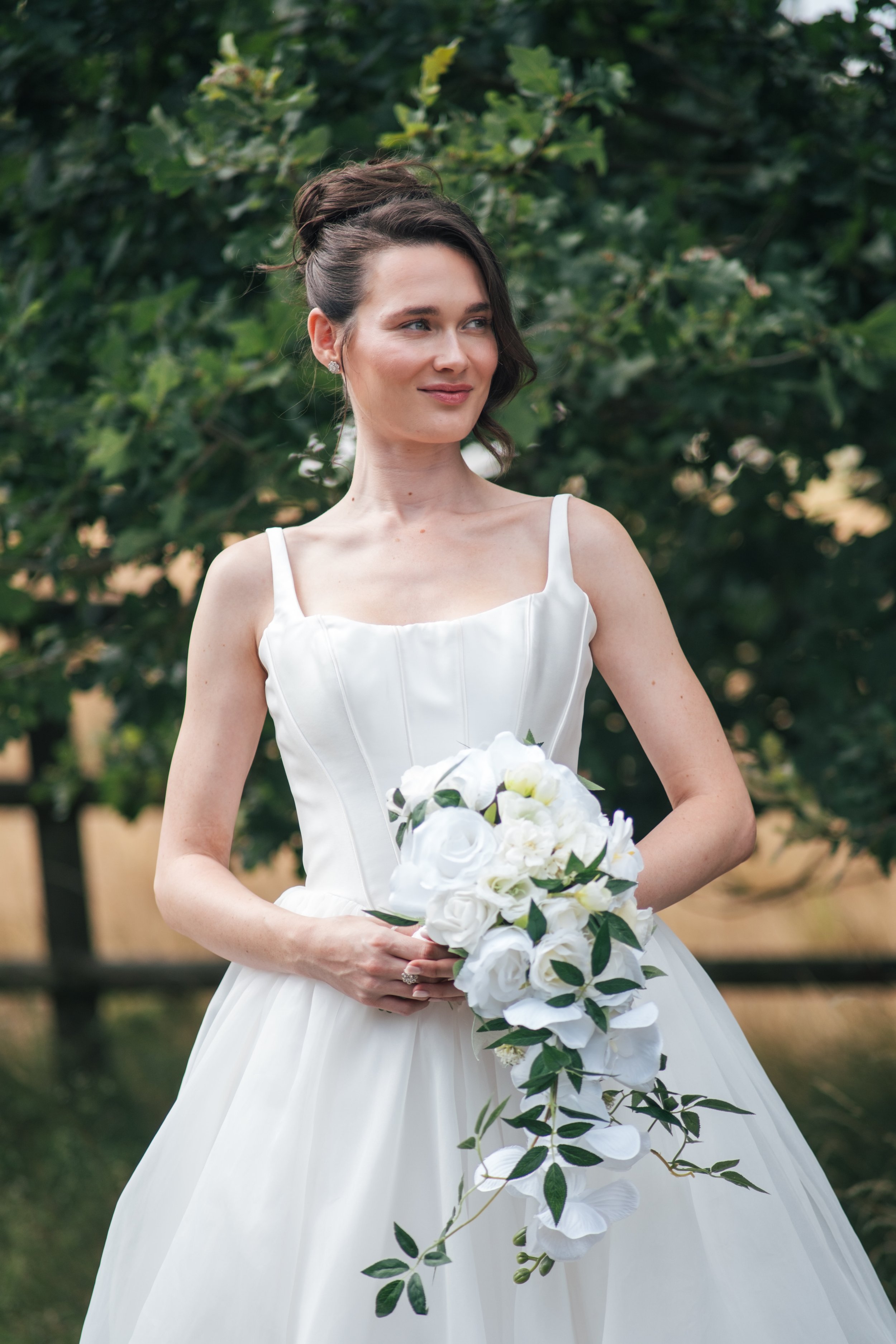 A woman in a white wedding dress holding a cascading bouquet of white flowers and green leaves, standing outdoors in front of green foliage.
