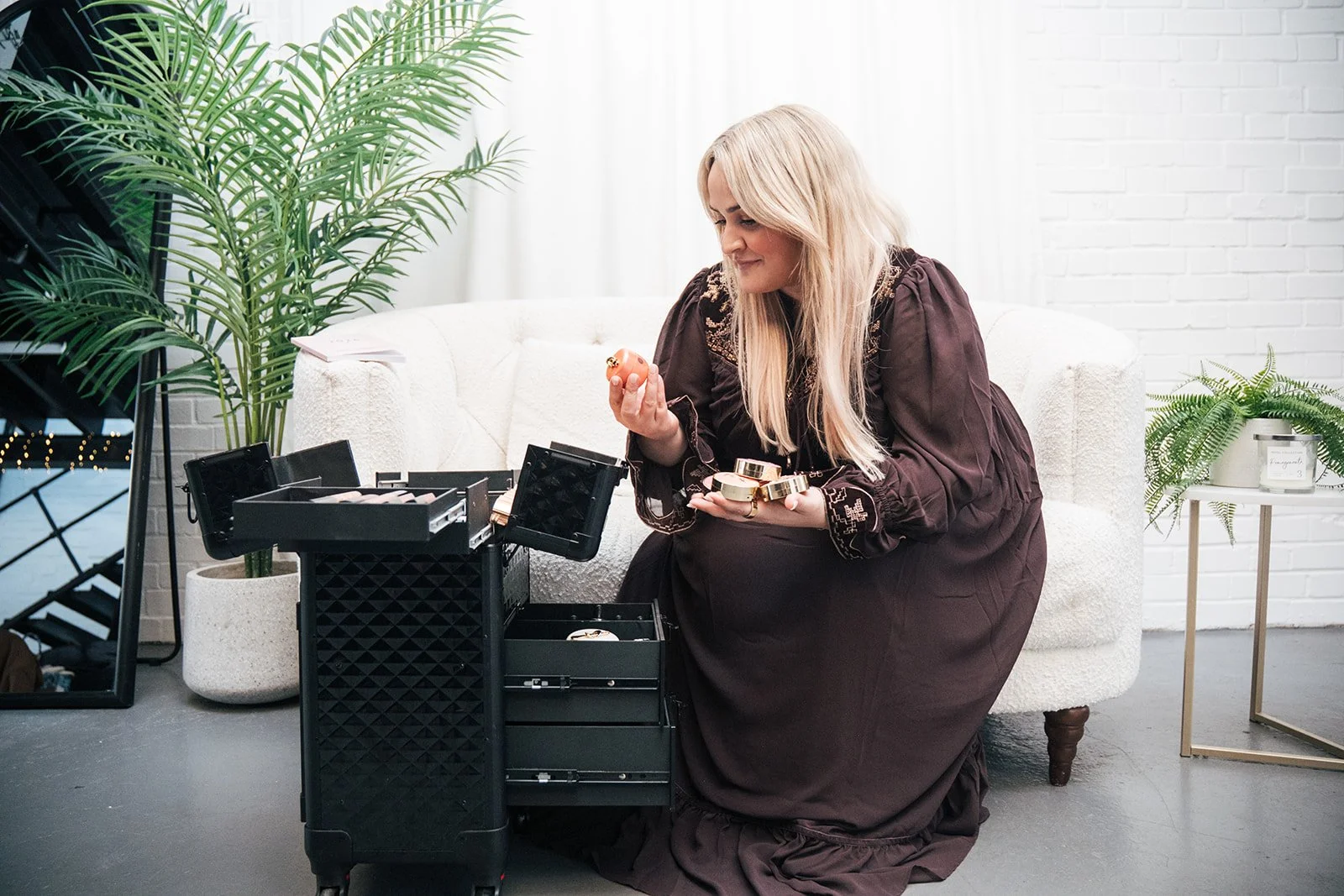 A woman with long blonde hair in a brown dress is sitting on a white couch, looking at beauty products in her hands. She is surrounded by jewellery boxes and a mirror, with large green plants nearby in a bright, modern room.