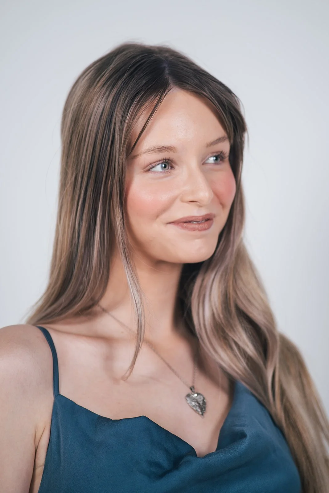 A young woman with long, light brown hair and blue eyes, wearing a navy blue top with thin straps and a silver heart-shaped pendant necklace, looking slightly to the side with a gentle smile.