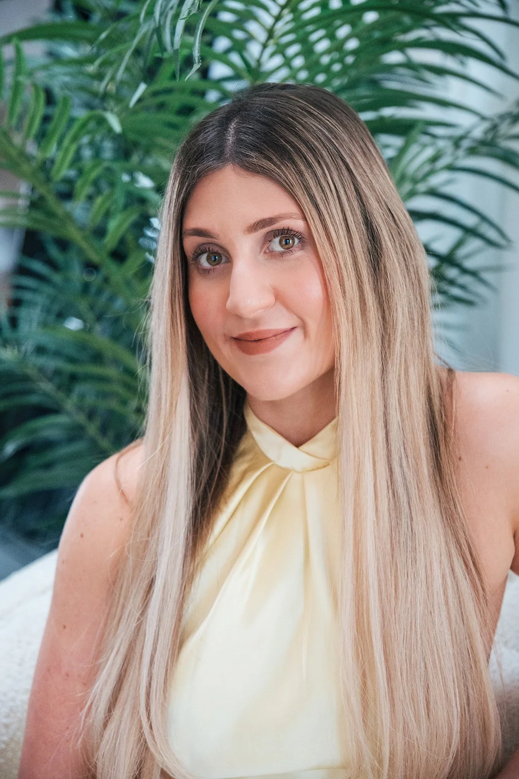 A woman with long blonde hair and light makeup poses in front of green indoor plants, wearing a light yellow sleeveless top.