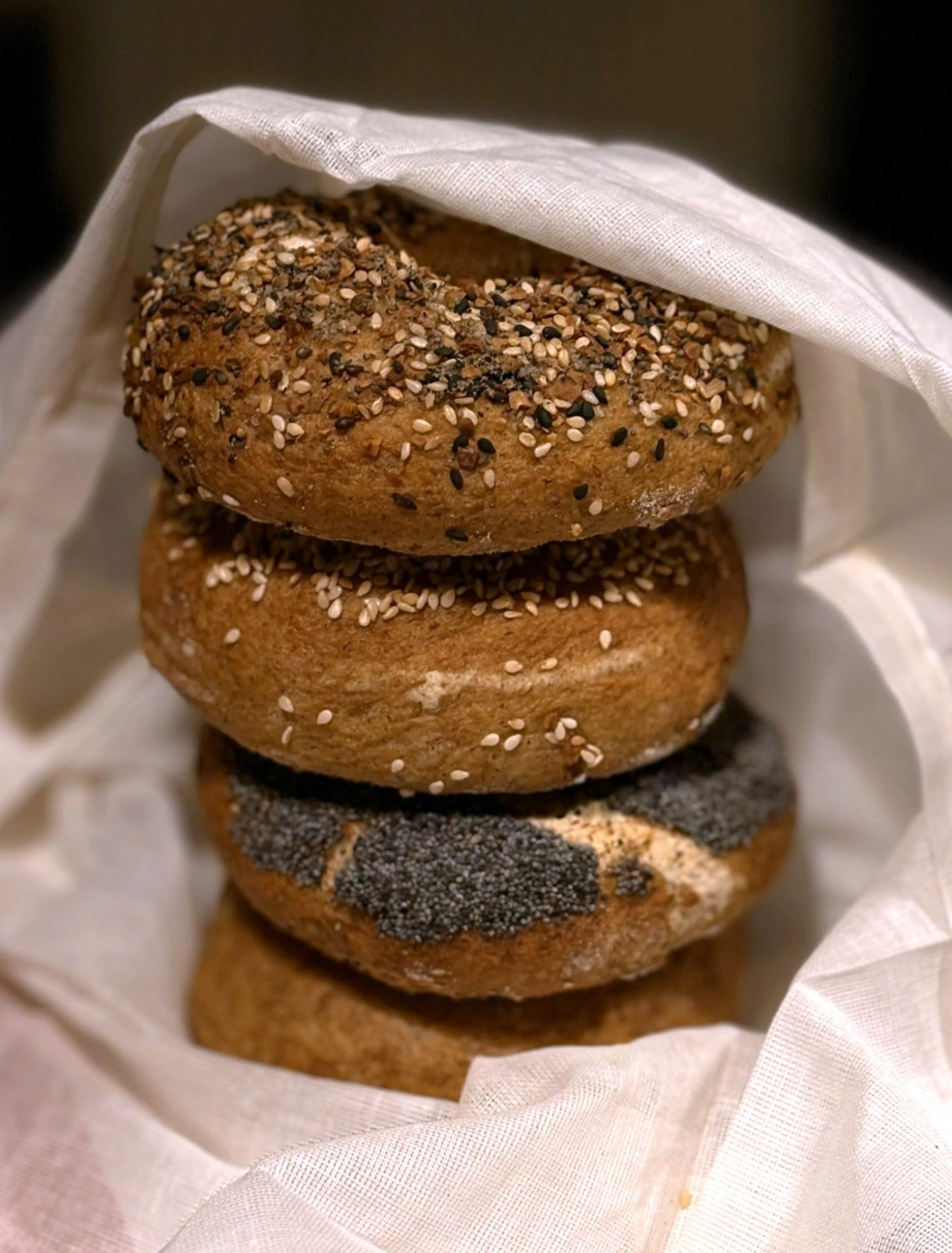 Assorted baked goods from The Common Crumb. Stacked bagels with toppings, including seeds and poppy seeds, wrapped in white cloth.