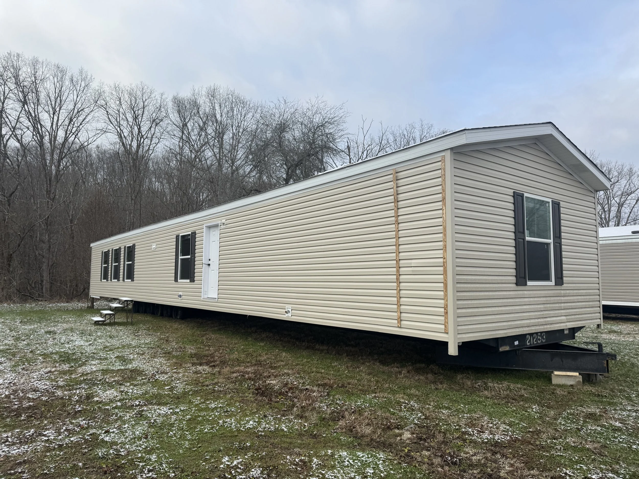 A beige mobile home with black shutters and windows, roller door entrance, and steps, situated on a slightly snow-dusted grassy lot with leafless trees in the background.