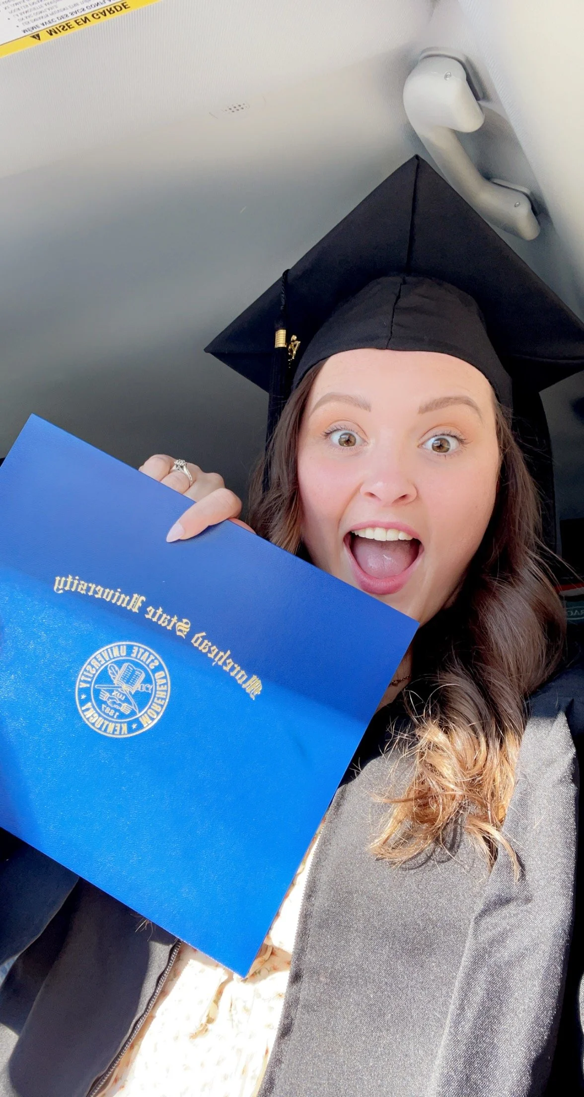 A woman wearing a graduation cap and gown holding a blue diploma cover with gold text and emblem, smiling with her mouth open, inside a vehicle. Morrison Marketing