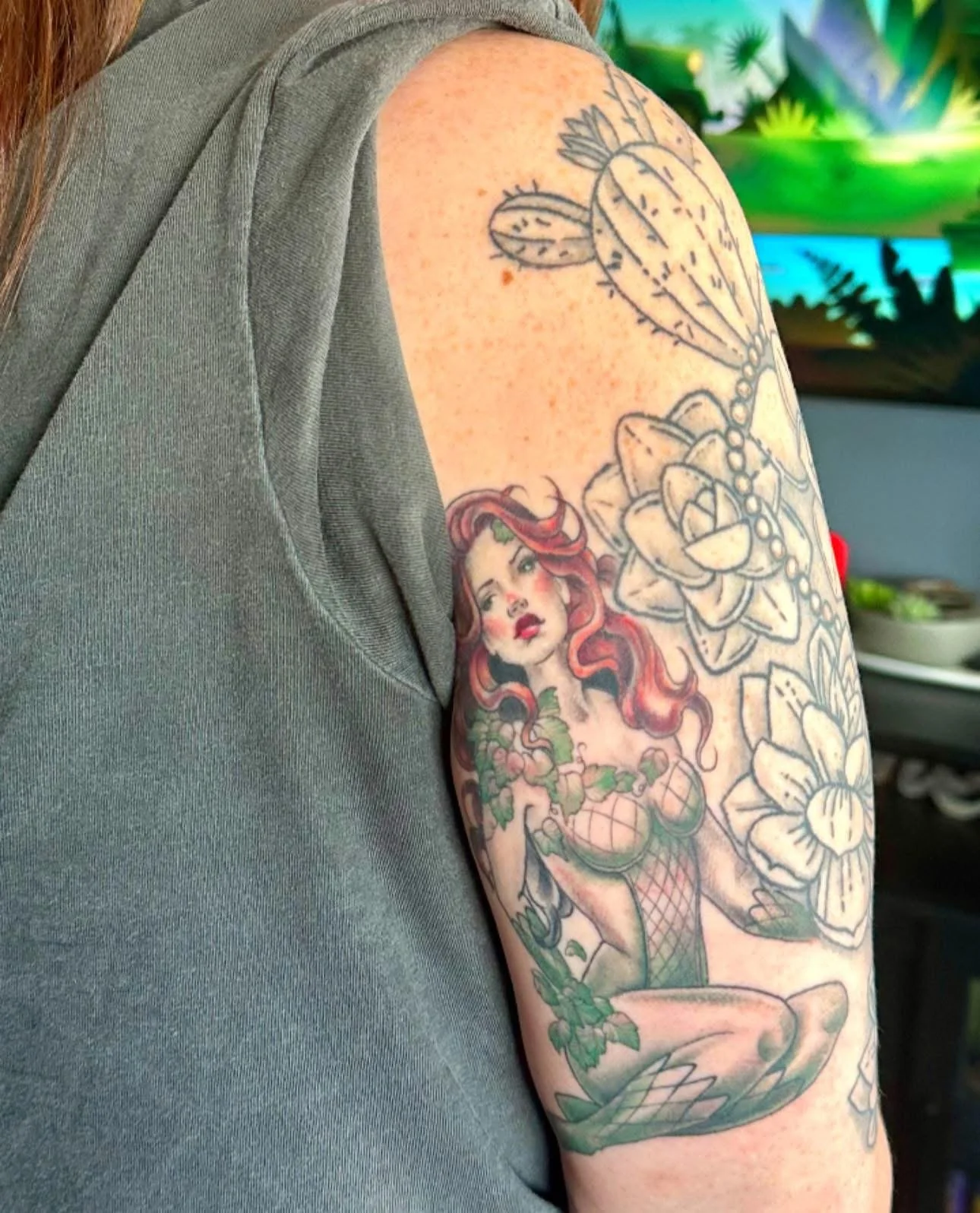 Tattoo of a mermaid with red hair, fishnet top, and a mermaid tail on the person's upper arm surrounded by large flowers and abstract shapes.