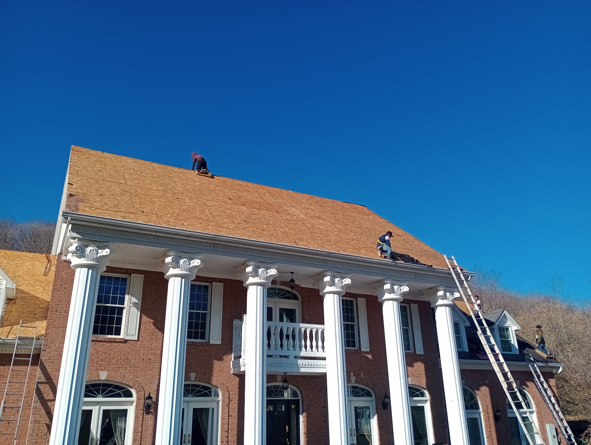 Workers installing a new roof on a large brick house with white columns, a small balcony, and multiple windows under a clear blue sky.