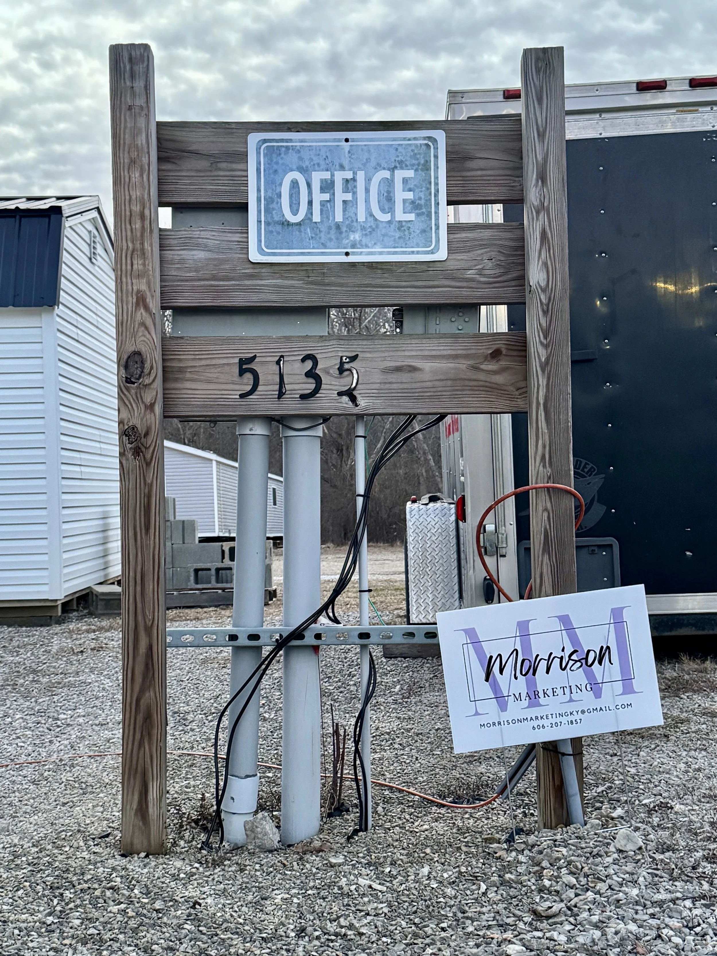 Wooden frame with a sign that says 'OFFICE' at the top, the number '5135' below it, and a business card for Morrison Marketing leaning against the right side of the frame. There are electrical wires and pipes at the base of the frame, and a trailer i