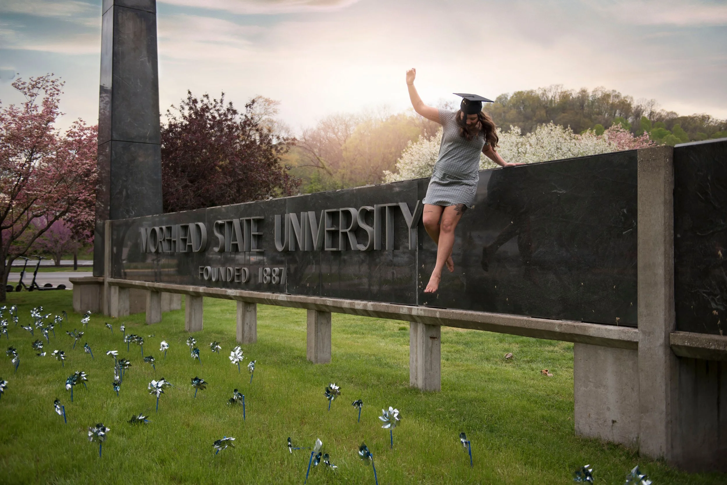 A woman in a graduation cap and dress is hanging off the edge of a large stone sign that reads "MOREHEAD STATE UNIVERSITY FOUNDED 1887". 