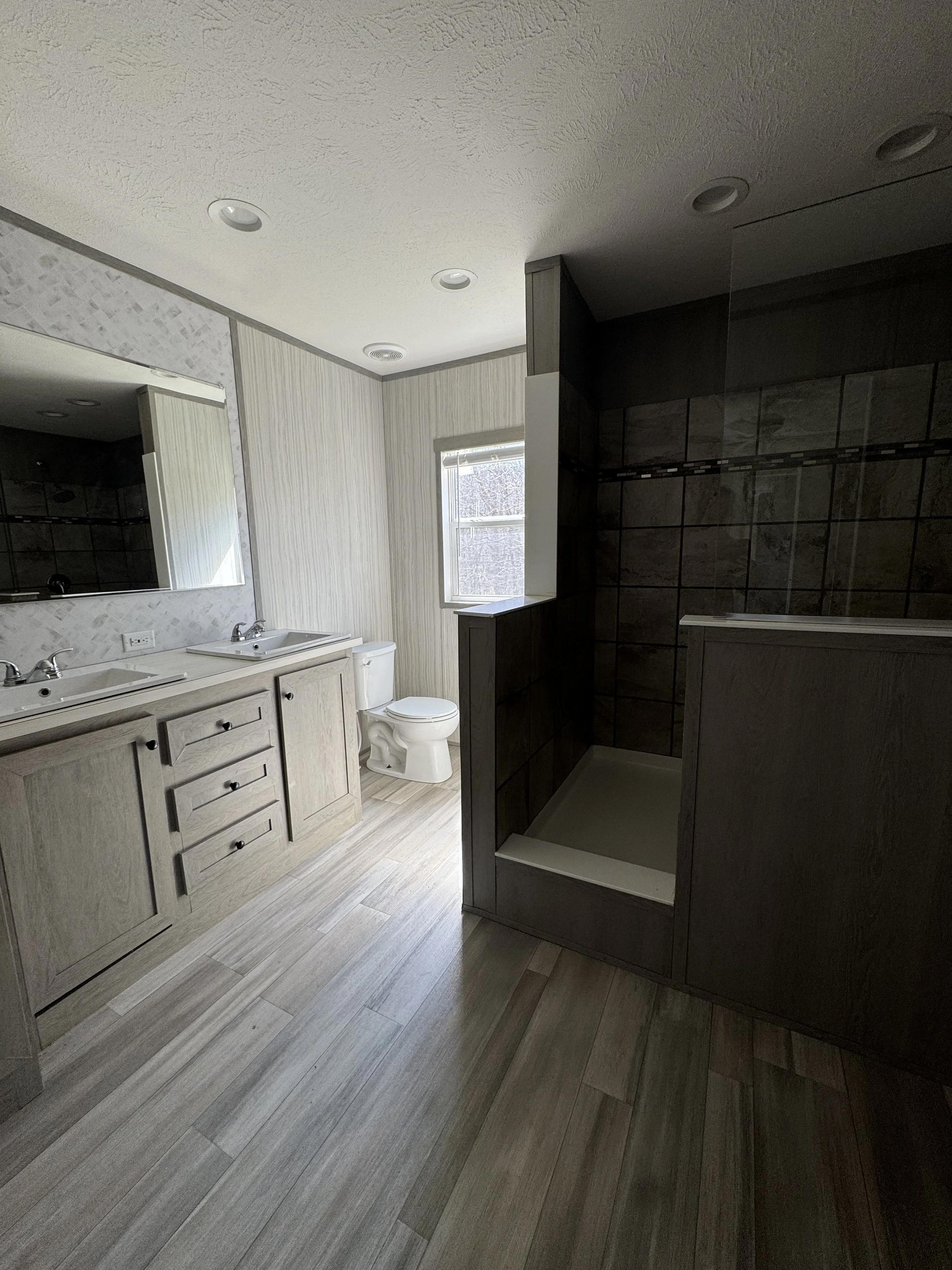 Bathroom with dual sinks, a toilet, a window, and a shower area with dark tiled walls.