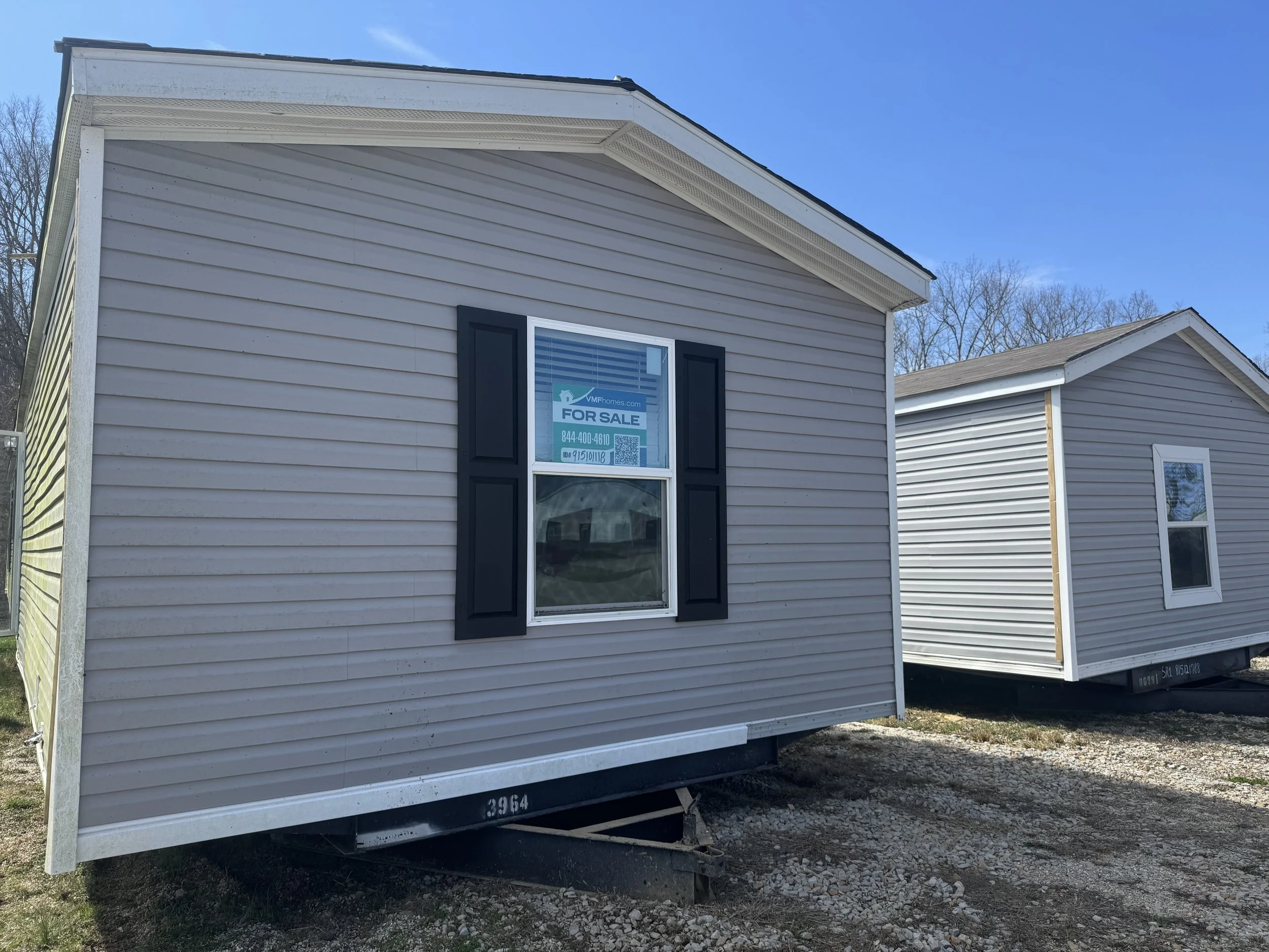 A beige mobile home with black shutters and a window with a 'For Sale' sign in it, situated on a gravel lot under a clear blue sky.
