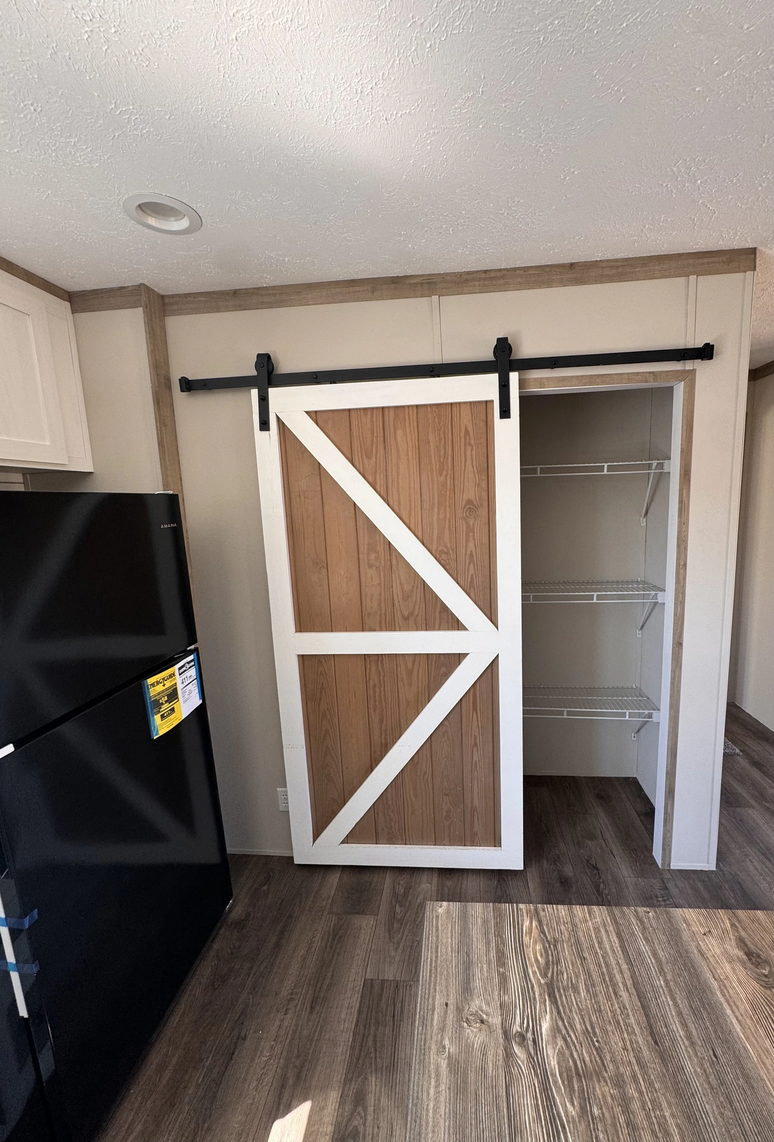 A partially assembled white and wood barn door on a black sliding hardware track next to an open closet with wire shelving within a room with wood flooring and beige walls.