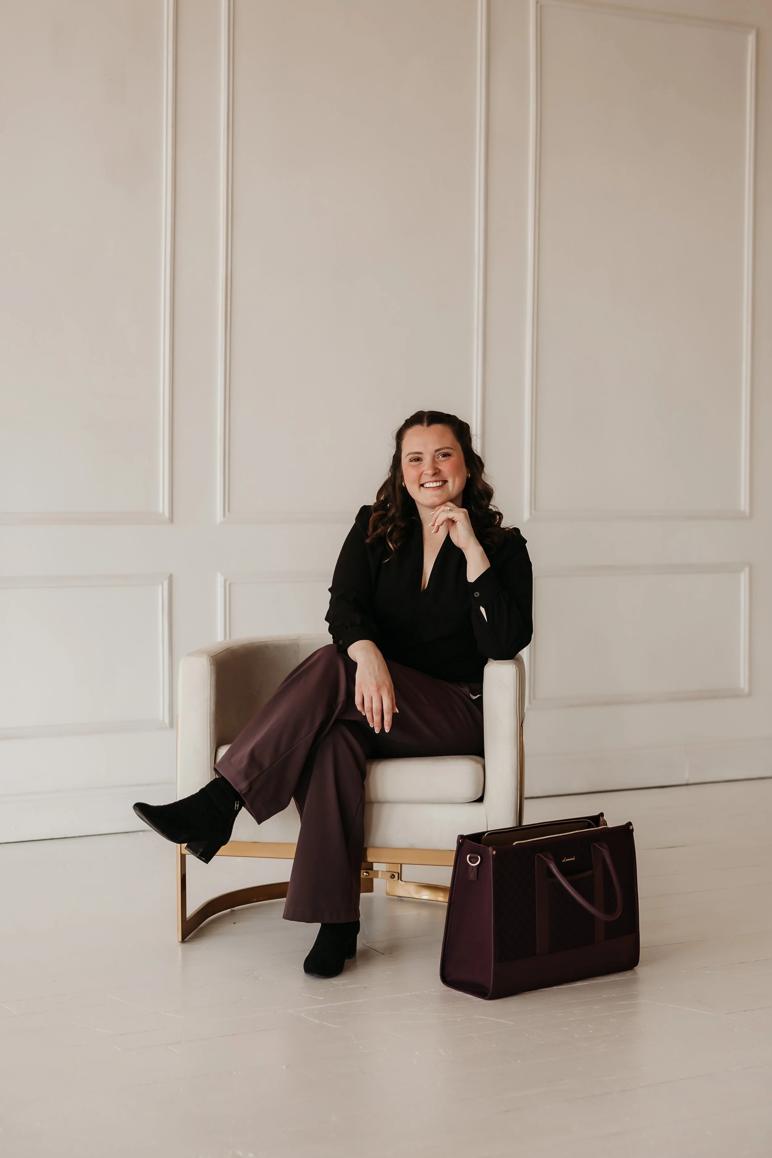 A woman sitting on a white chair, smiling, wearing a black top, dark pants, and black boots, with a large purple handbag on the floor beside her in a minimalist white room.