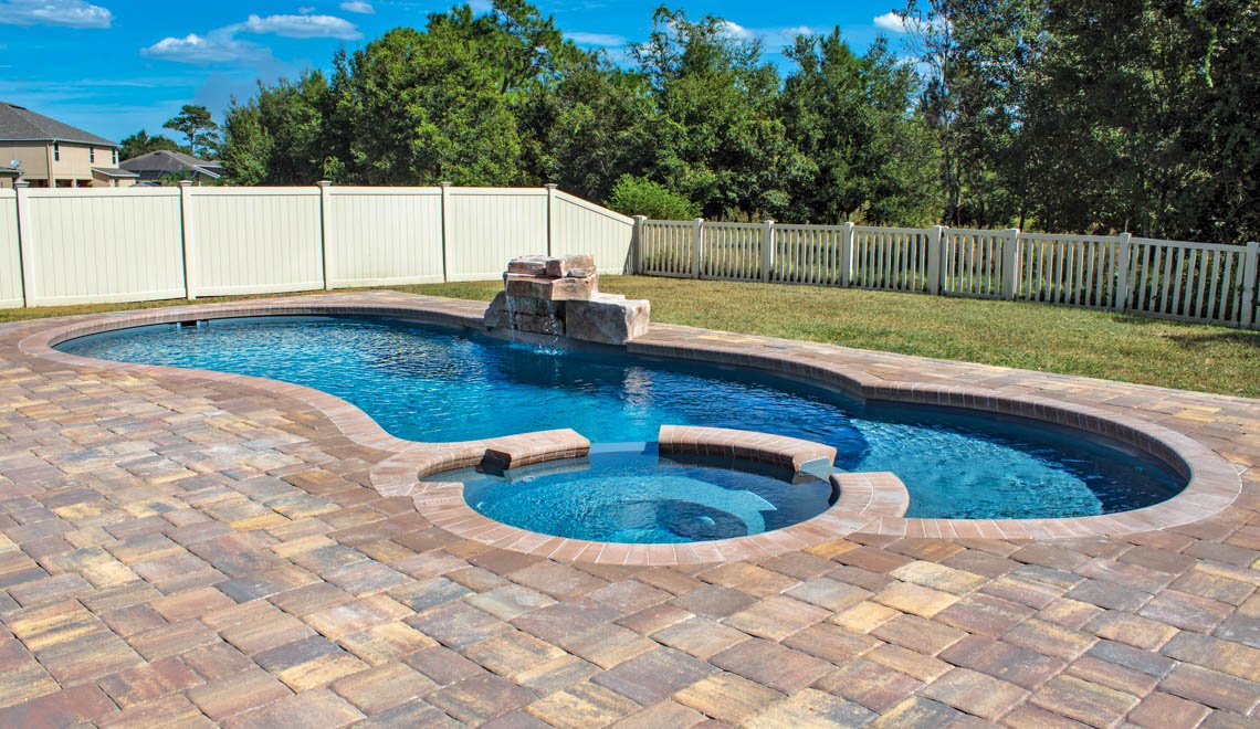 Backyard swimming pool with a hot tub, brick deck, white fence, green grass, and trees in the background under a blue sky.