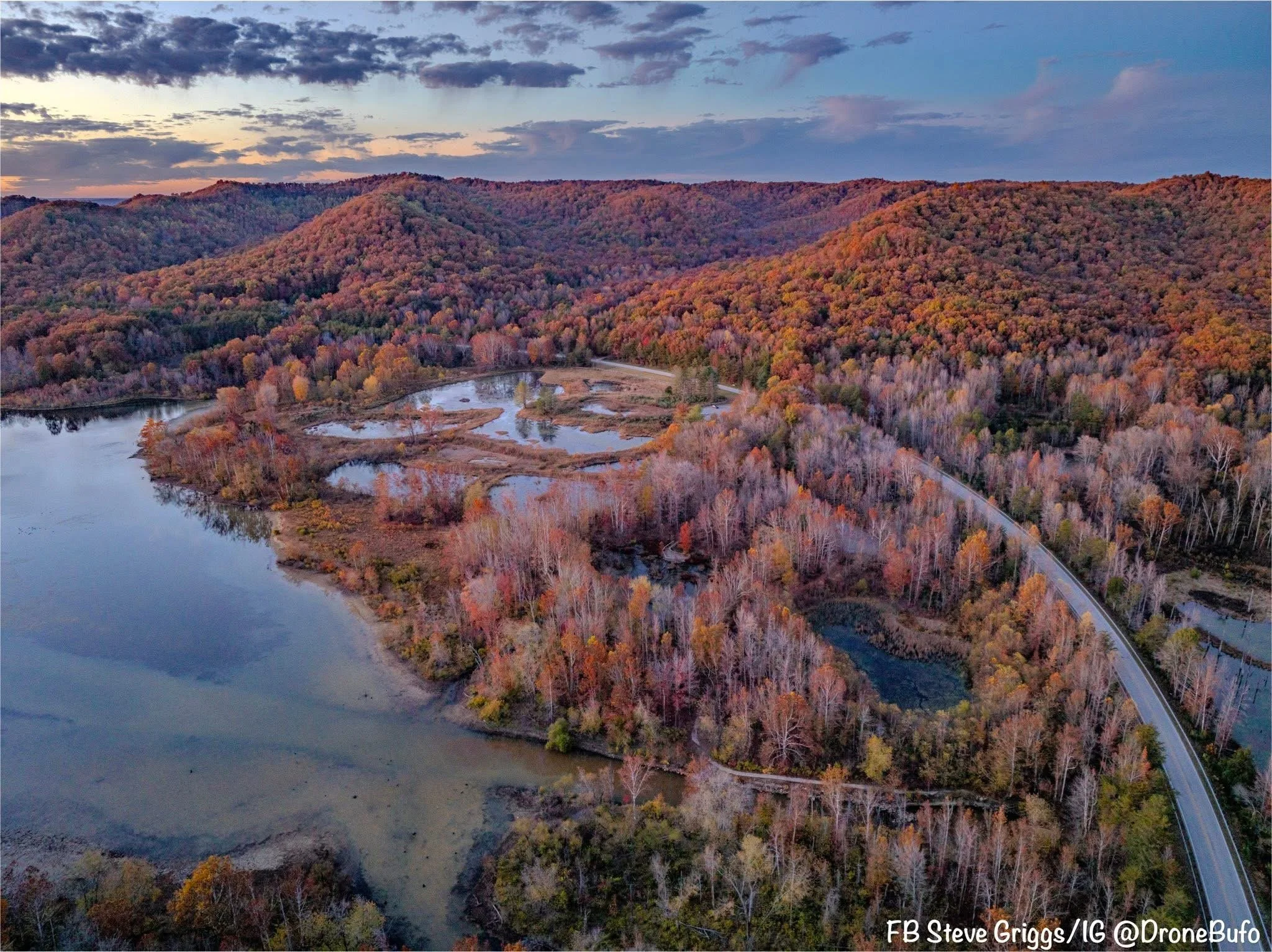 An aerial view of a landscape with a winding river, wetlands, and forested hills covered in autumn foliage during sunset.