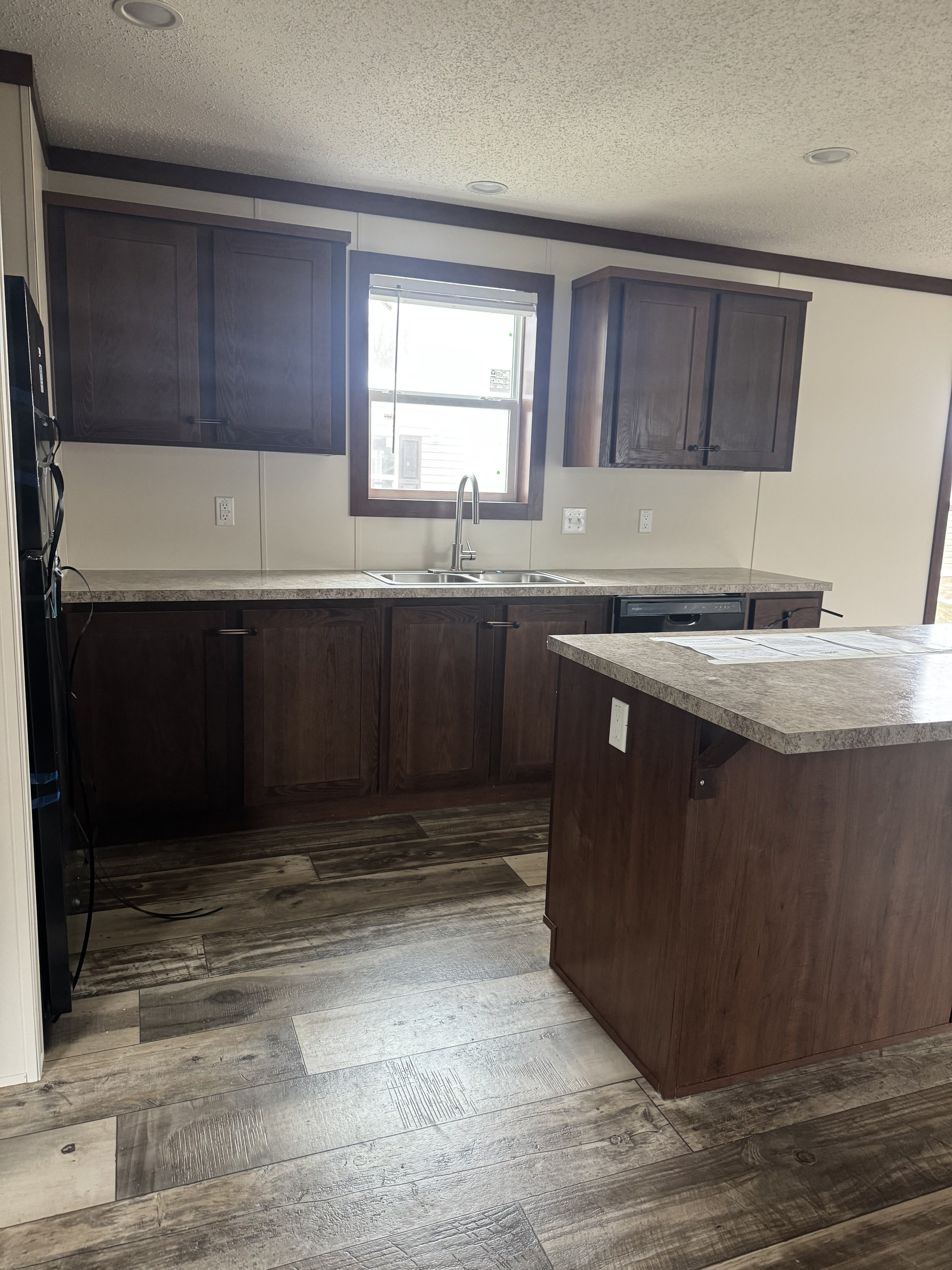 Empty kitchen with dark wood cabinets, beige countertops, a double sink under a window, and wood flooring.