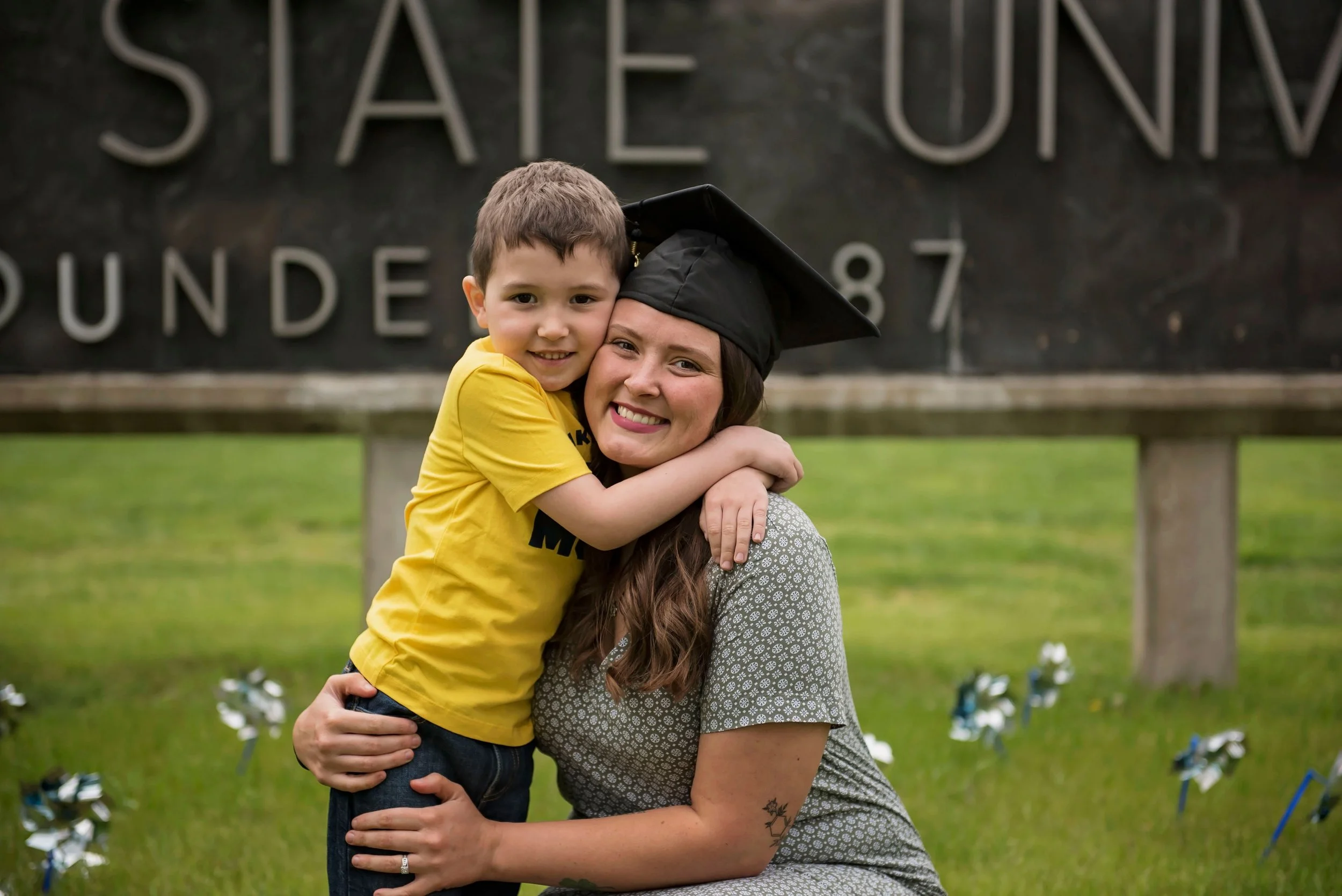 A woman in a graduation cap and gown hugging a young boy in a yellow shirt at a graduation ceremony.