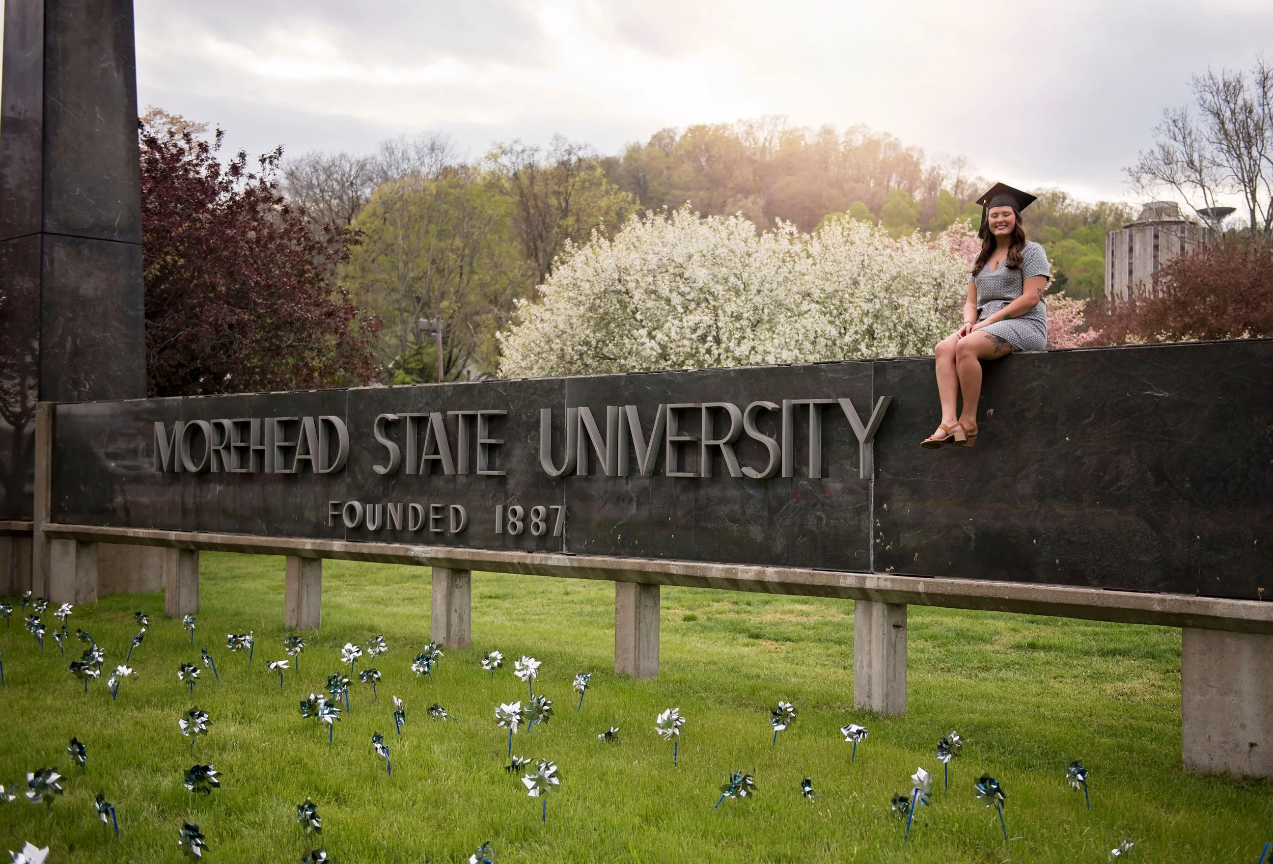 A woman in a graduation cap and dress sitting on a stone sign reading 'Morehead State University Founded 1887' with blooming trees and a cloudy sky in the background.