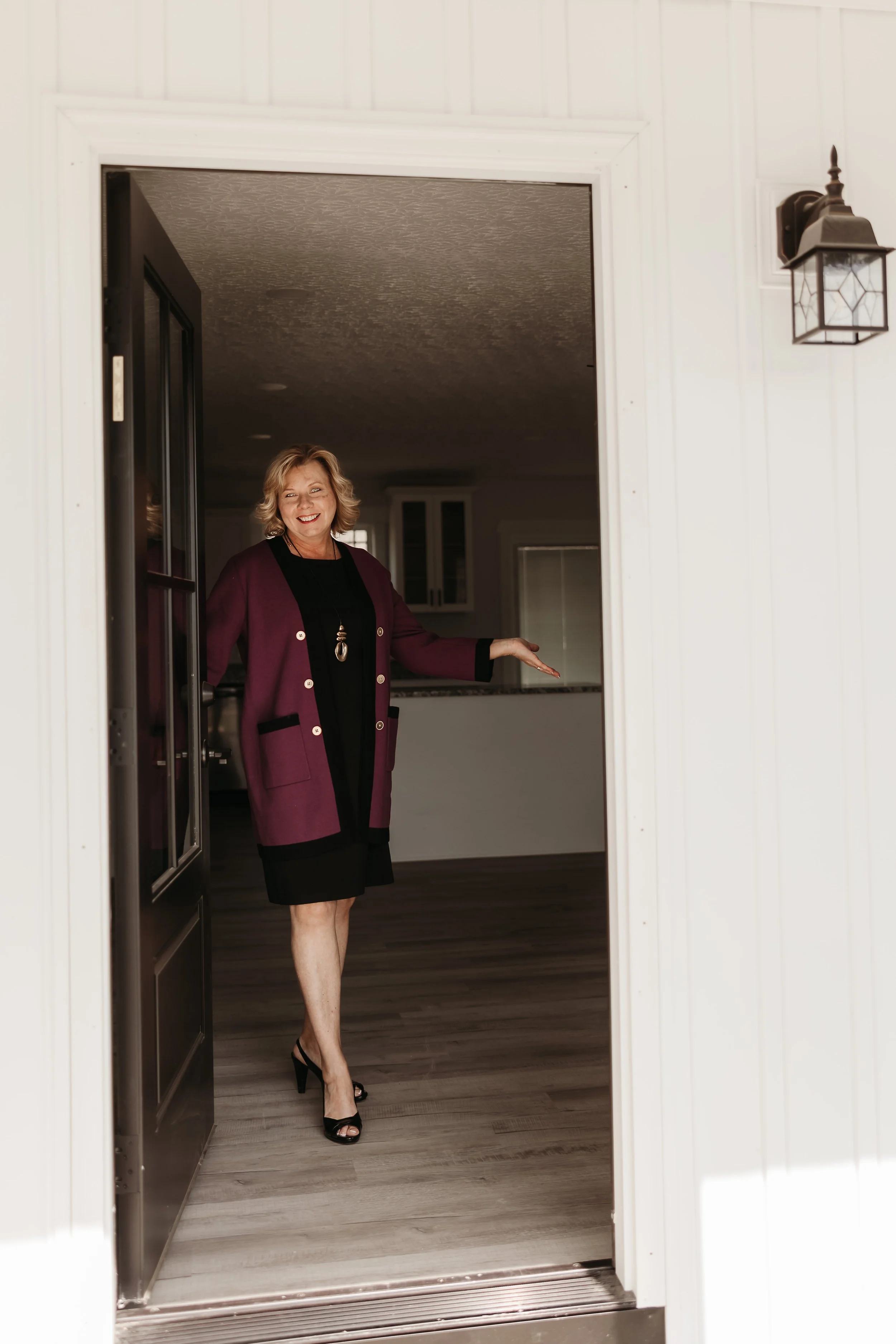 A woman with blonde hair wearing a purple blazer, black dress, and black high heels standing at the door of a house, smiling and welcoming.