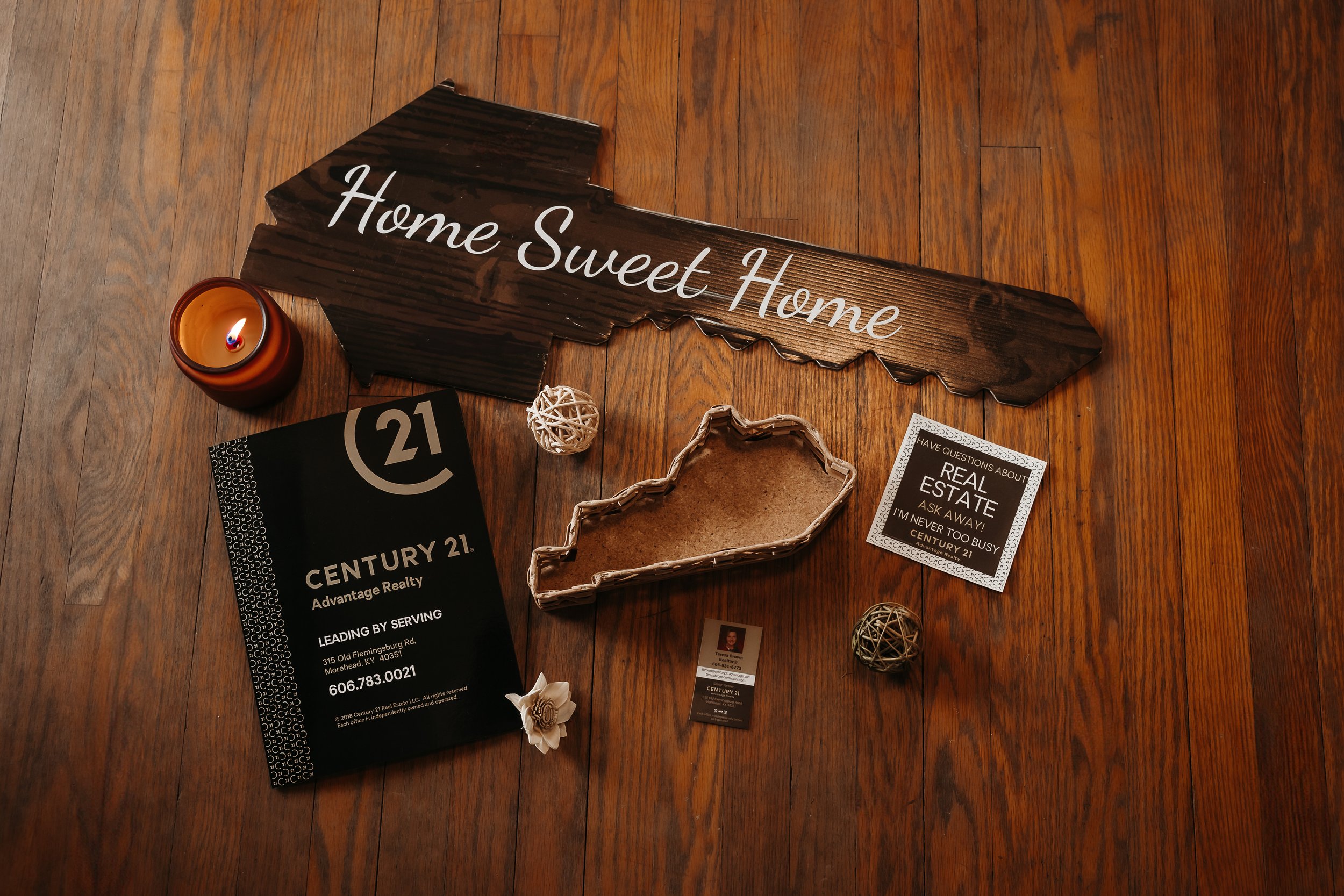 A wooden sign with the words 'Home Sweet Home' on a hardwood floor. Surrounding the sign are a lit candle, a black Century 21 real estate brochure, a small white decorative ball, a cardboard heart-shaped tray, a Century 21 business card, a small whit