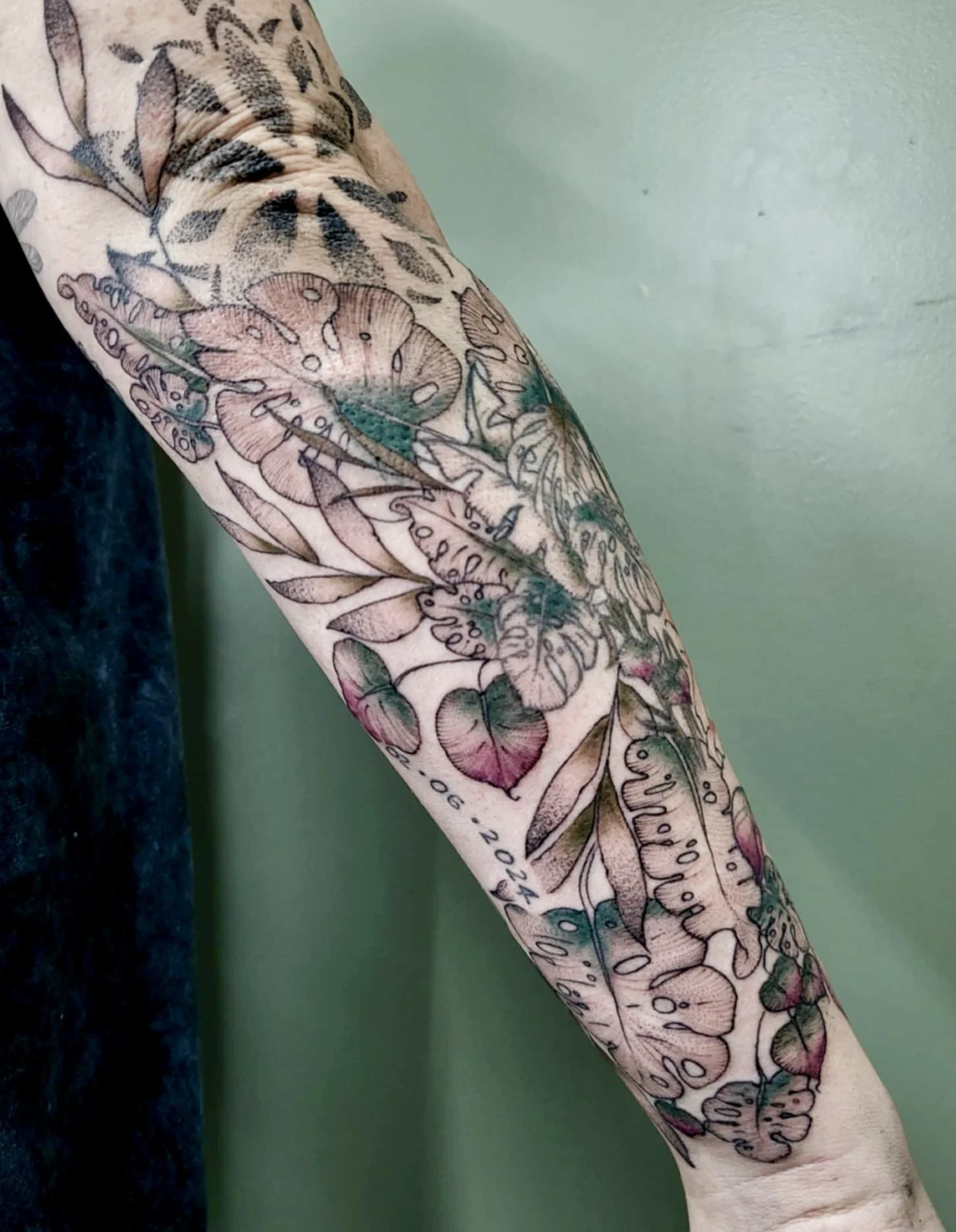 Tattoo sleeve featuring floral and leafy designs, some colored in pink and green, with the date 06.2024.