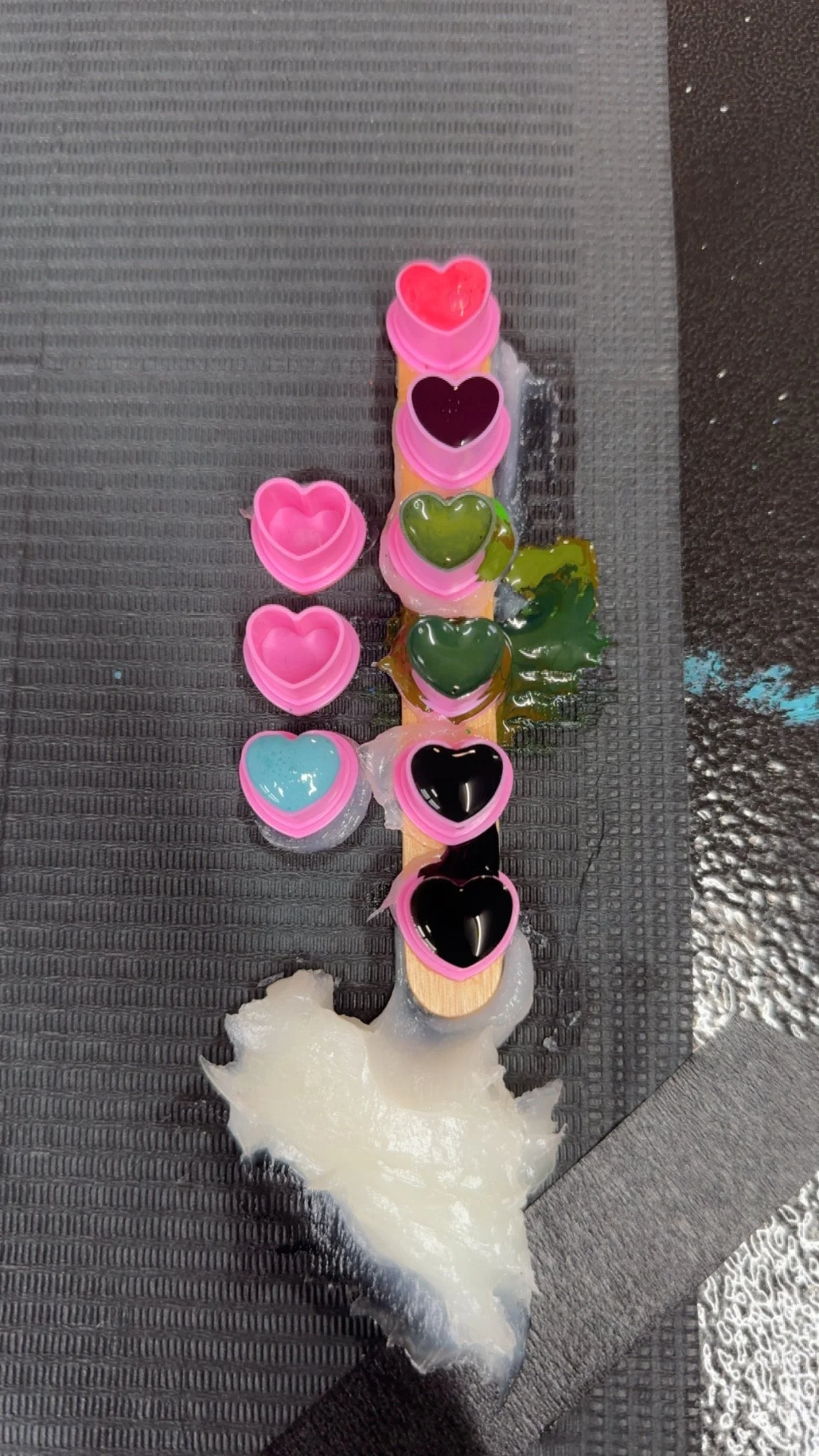 Assorted colorful gummy candies in shape of hearts, some with pink edges, on a wooden stick, with melted white candy on a black textured surface.