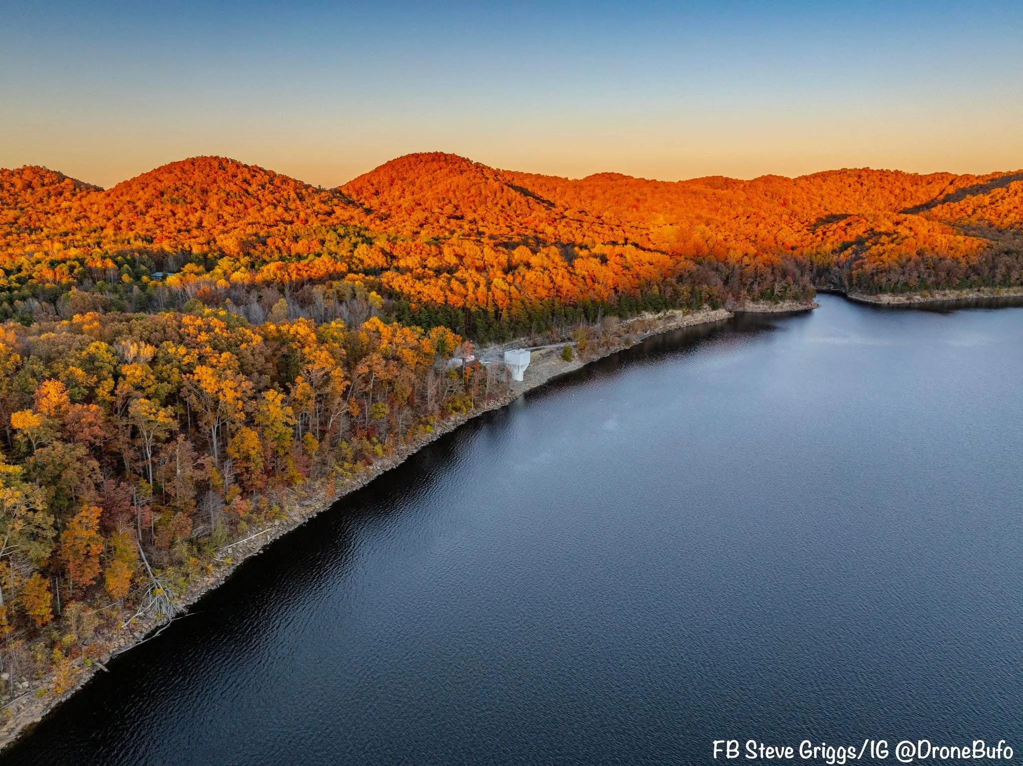An aerial view of a lake edged by a forested shoreline with trees displaying fall colors and hills covered with orange foliage in the background during sunset.