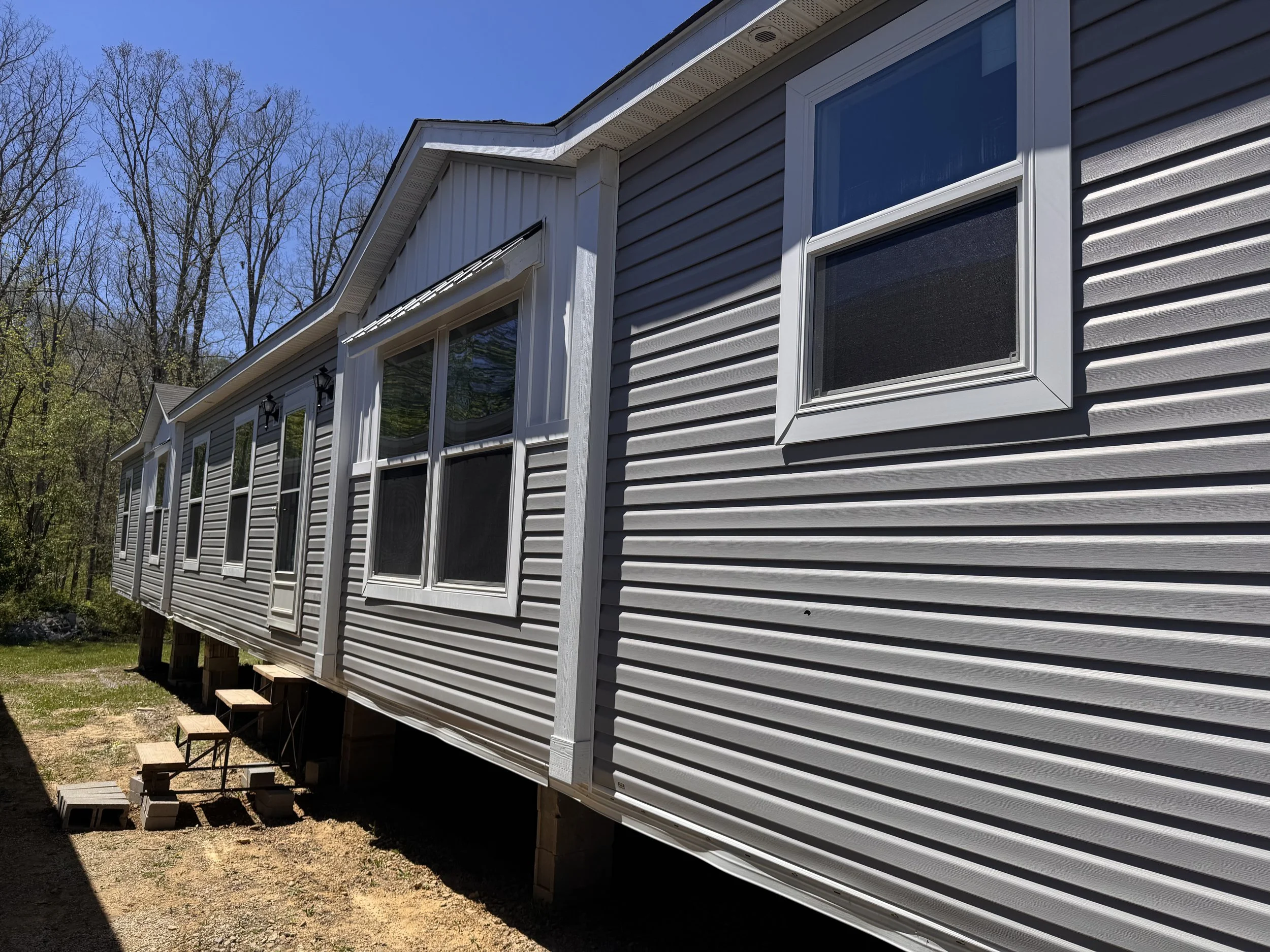 Side view of a house with gray vinyl siding, multiple white-framed windows, and a small staircase leading to the entrance, set against a background of leafless trees and a clear blue sky.