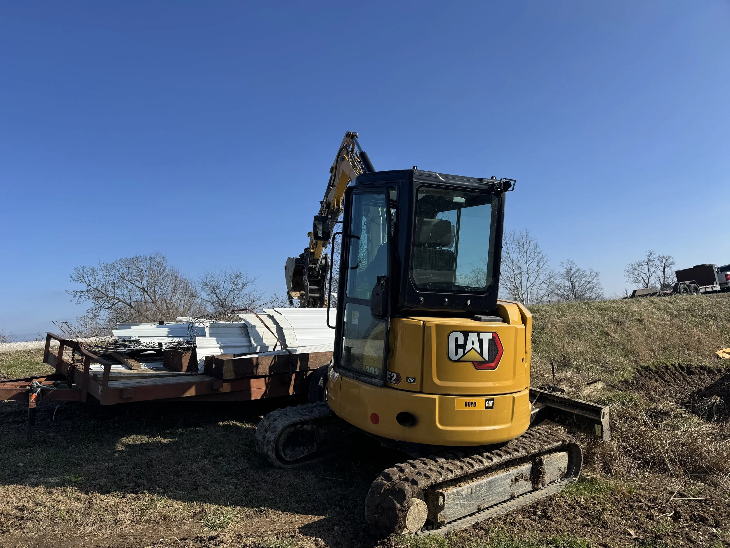 A yellow Caterpillar mini excavator with a black cab, working on a construction site with a grassy hill and trees in the background under a clear blue sky. It is positioned on a patch of dirt and is attached to a flatbed trailer with white metal beam